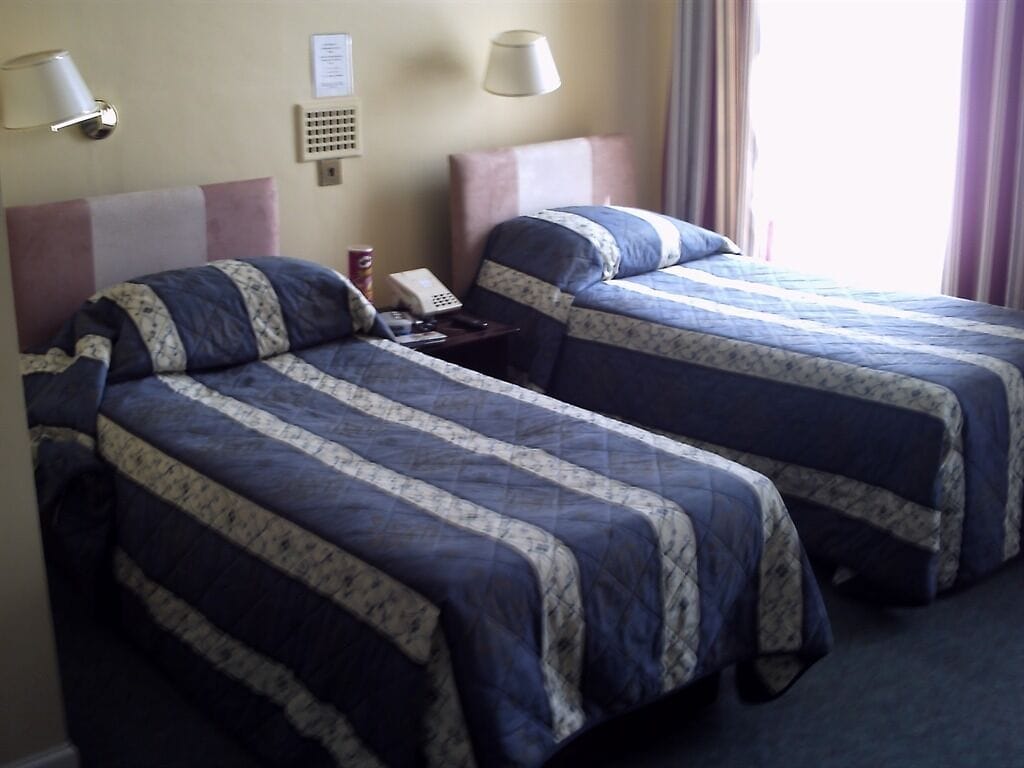 Ein Hotelzimmer mit zwei Betten, jeweils mit einem blau-weißen Musterbezug, einem Nachttisch mit Lampe und einem an der Wand montierten Telefon.