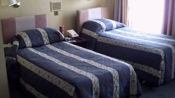 A hotel room with two beds, each with a blue and white patterned bedspread, a bedside table with a lamp, and a wall-mounted phone.