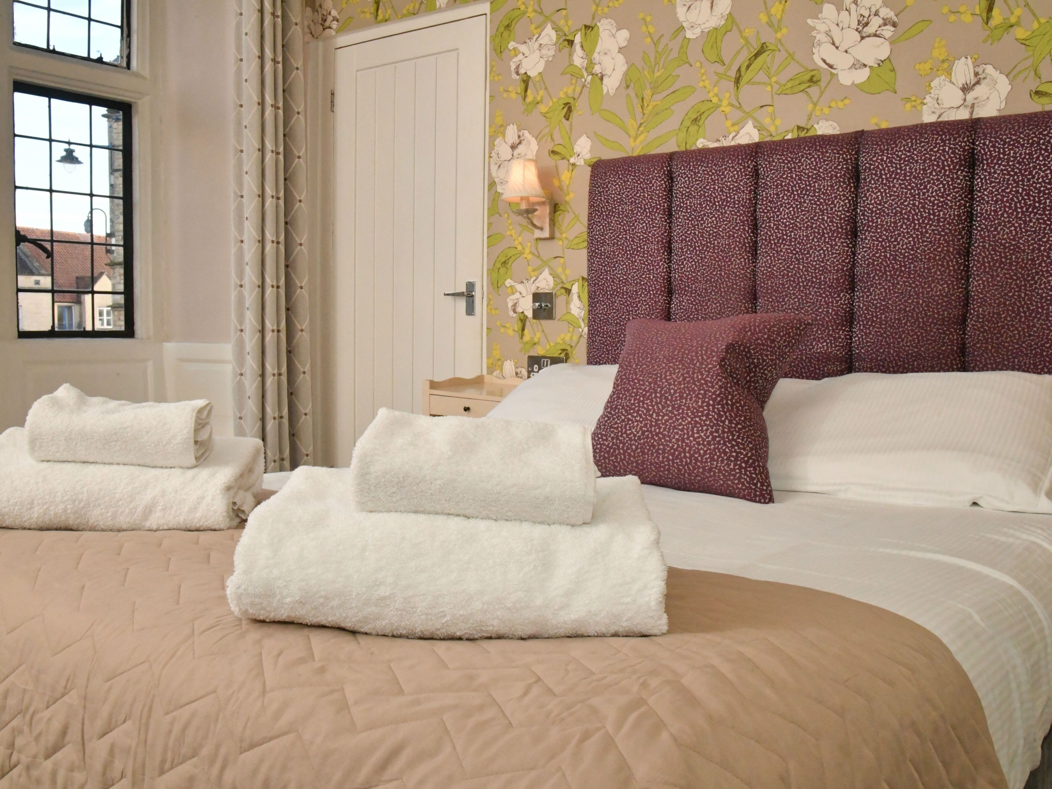 A neatly made bed with white and purple pillows, a beige quilted bedspread, and two folded white towels on the bed.