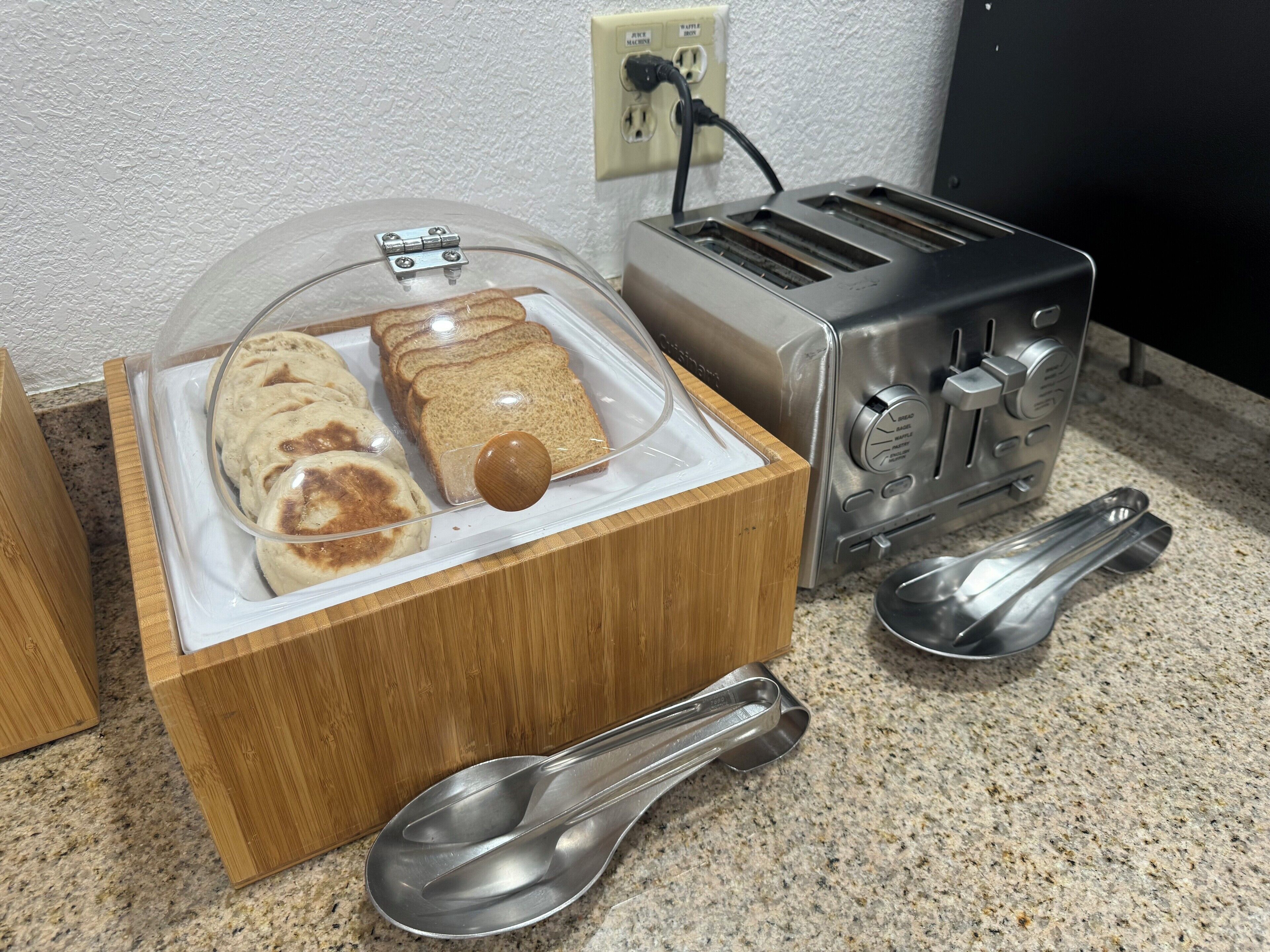 Una cocina con una caja de pan, tostadora y dos cucharas de metal sobre una encimera de granito.