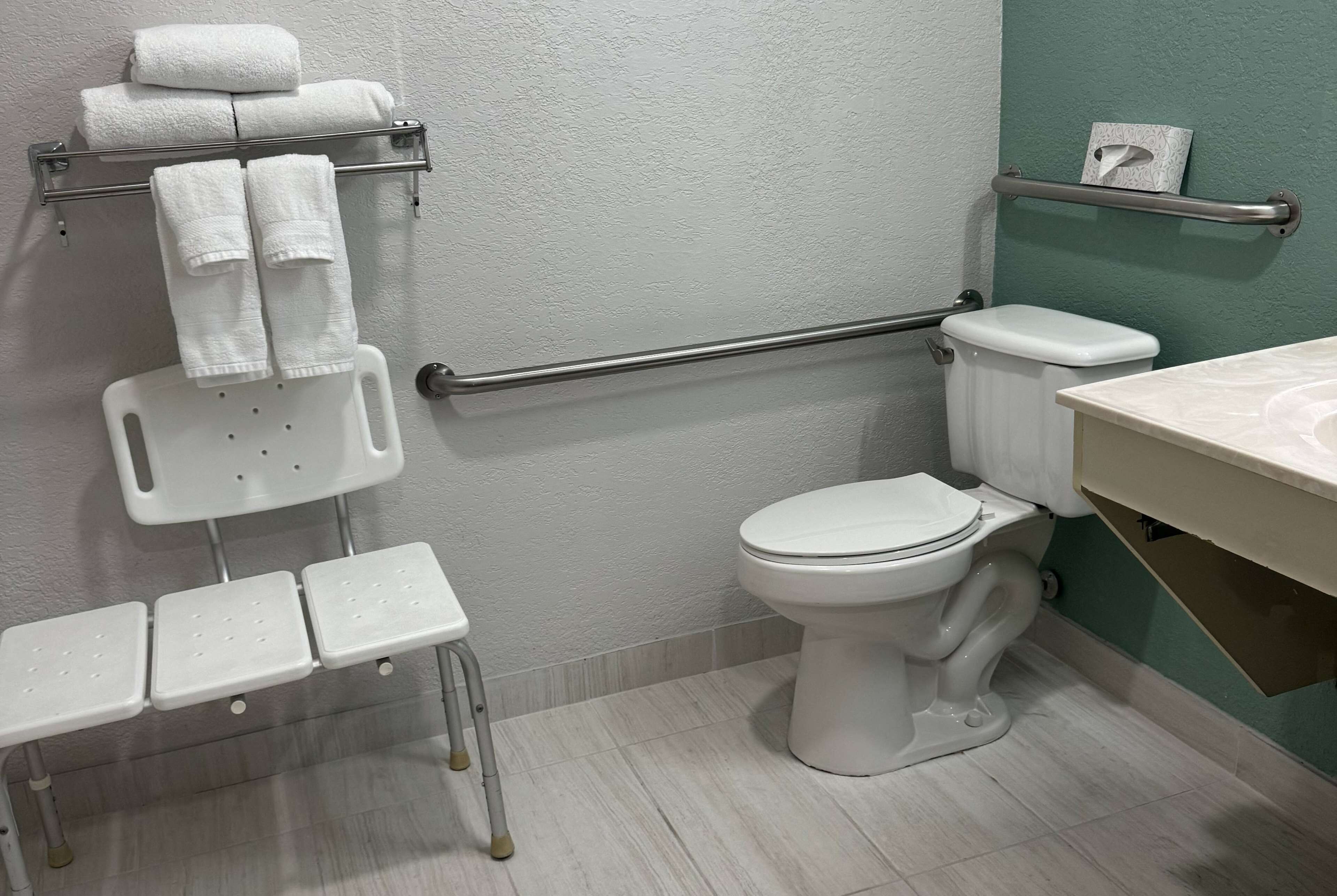 A bathroom with a white toilet, a white sink, a grab bar, a white bench, and a towel rack with towels.