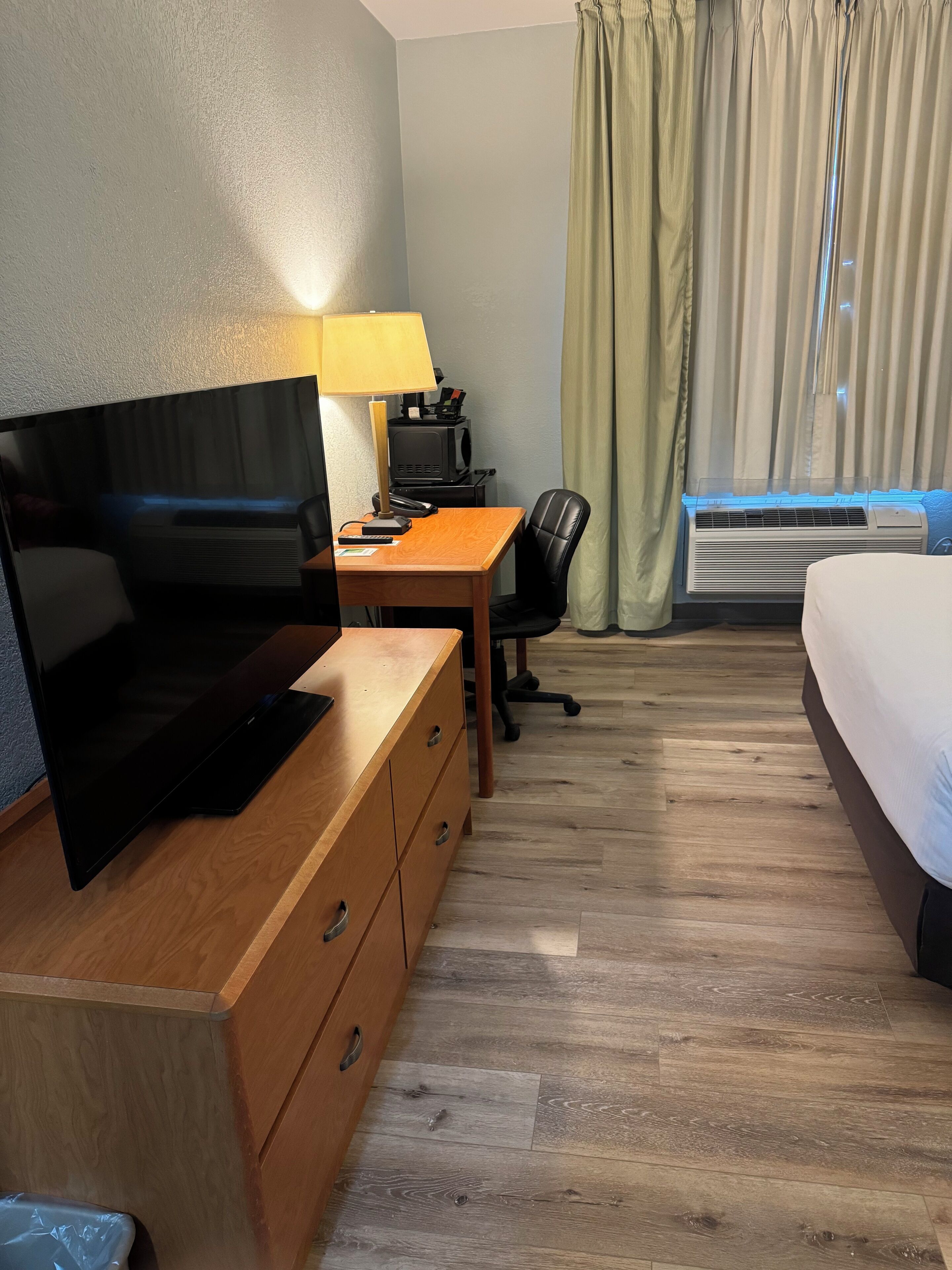 A hotel room with a wooden desk, a television, a lamp, a bed, and a window with curtains.