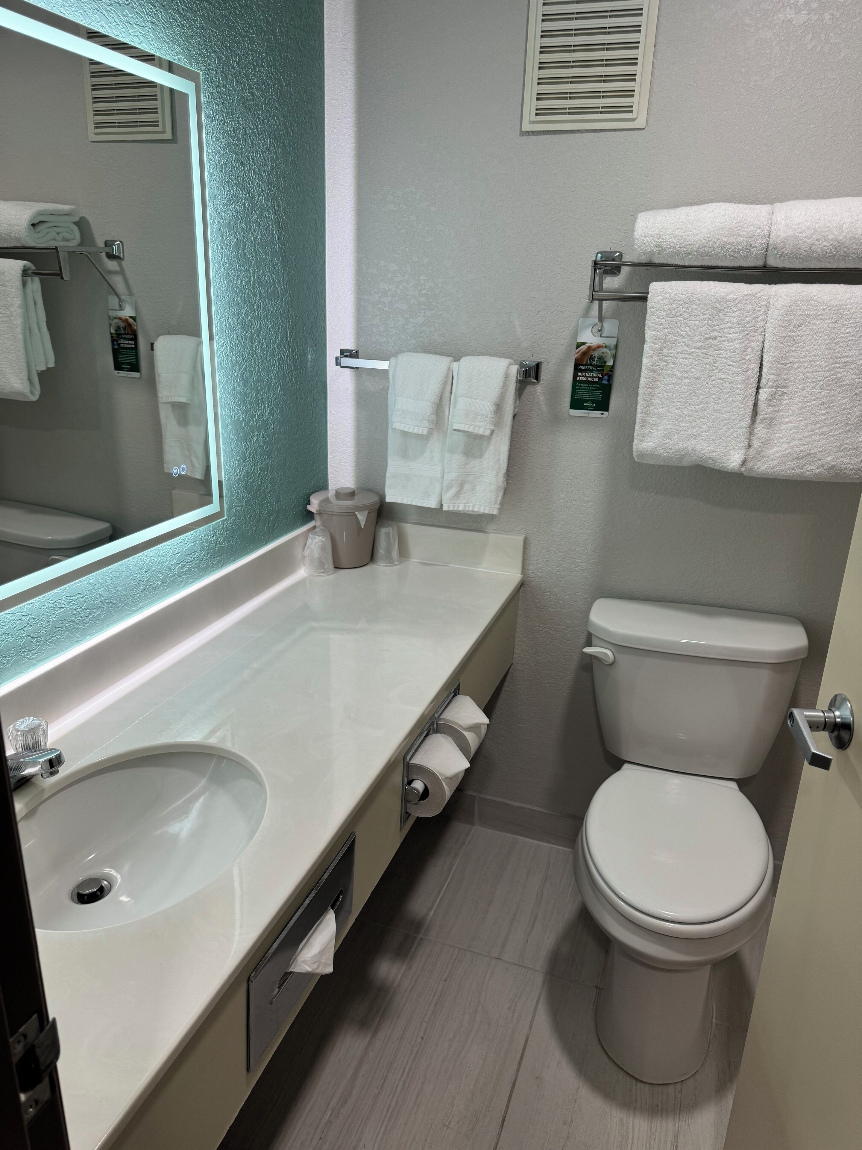 Un baño con lavamanos blanco, inodoro y espejo. Hay toallas colgadas en un perchero y un dispensador de toallas de papel.