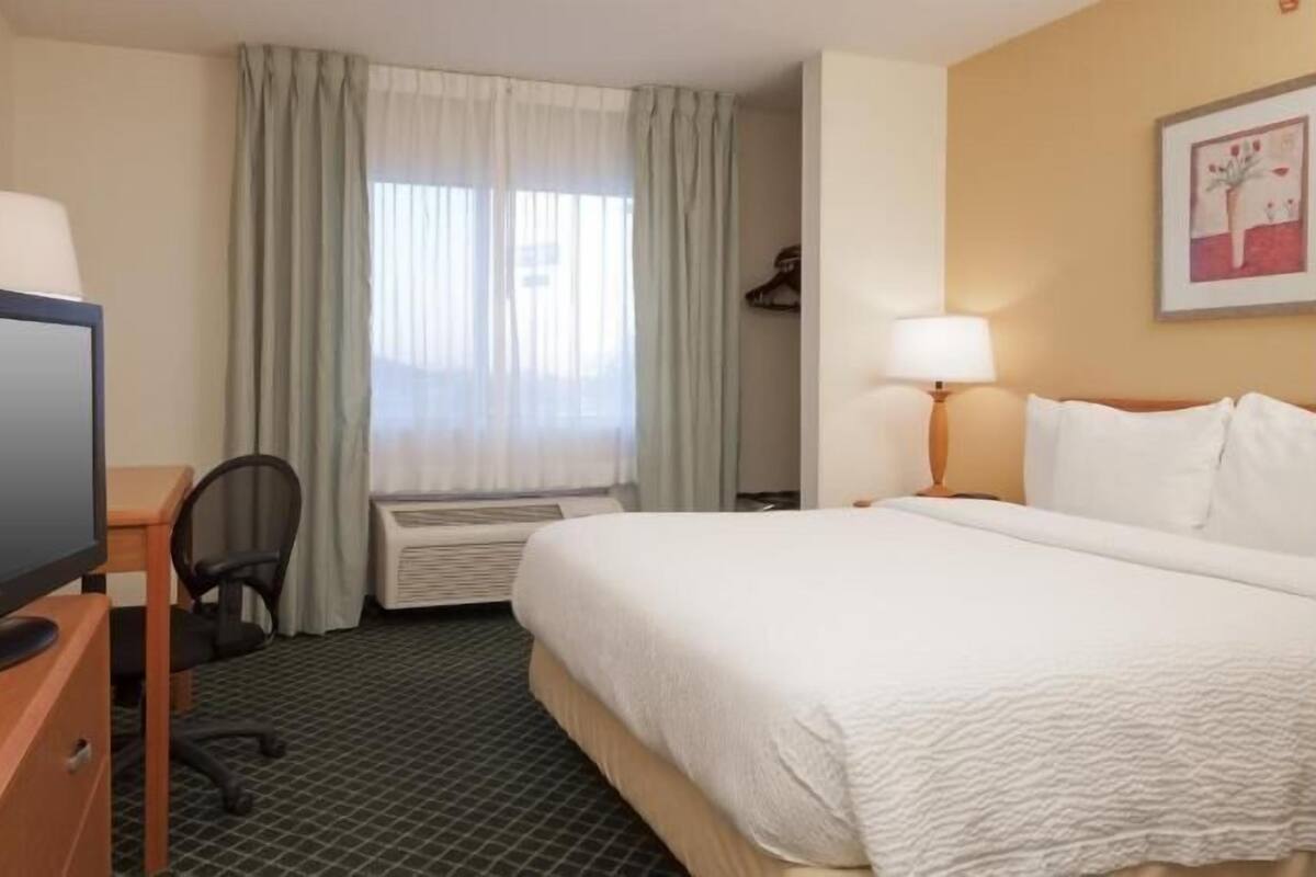 Habitación de hotel con cama, escritorio con computadora, televisión, ventana con cortinas y lámpara en la pared.