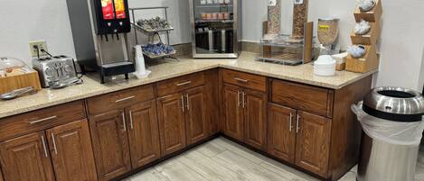 A corner kitchen with a coffee machine, toaster, refrigerator, and various food dispensers.