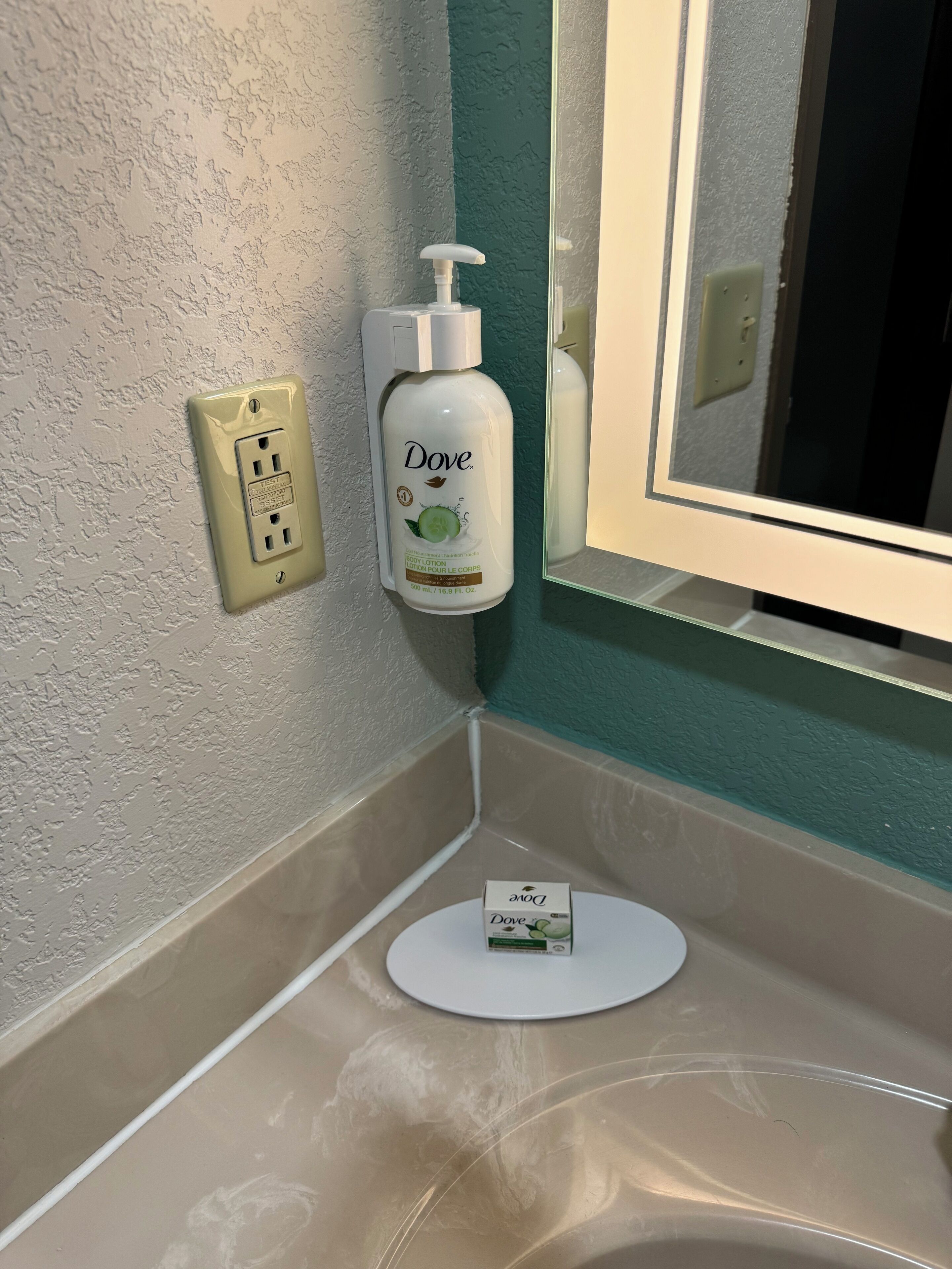 A bathroom with a Dove body lotion dispenser mounted on the wall, a white sink, and a mirror.