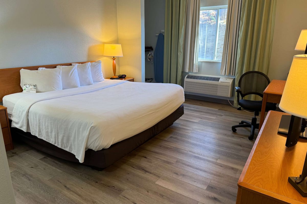 Habitación de hotel con una cama grande, un escritorio, una silla, una lámpara, una tabla de planchar y un ventanal con cortinas.