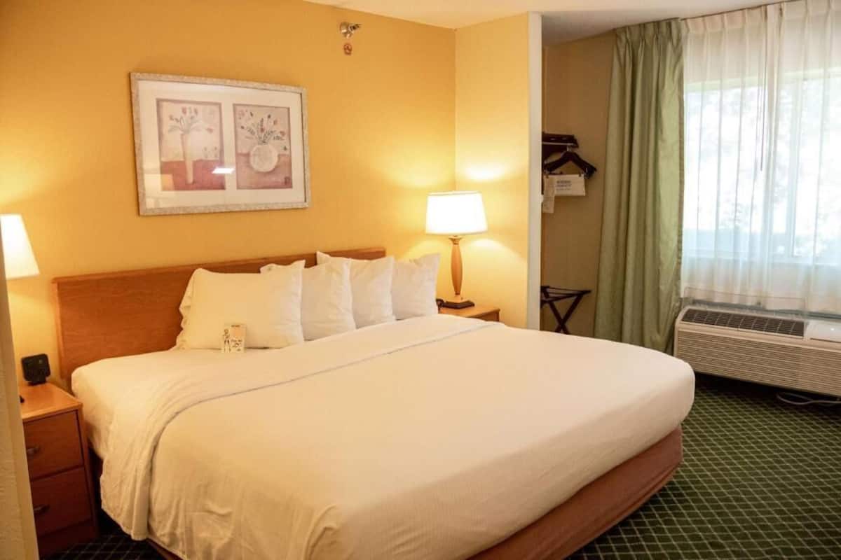 Una habitación de hotel con una cama grande, una mesita de noche con lámpara, un cuadro enmarcado en la pared y una ventana con cortinas.