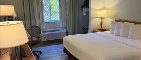 A hotel room with a bed, two bedside lamps, a desk with a lamp, a chair, a window with curtains, and a wall-mounted heater.