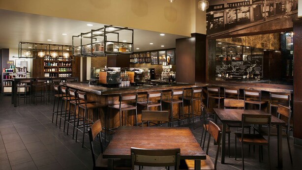 A bar area with wooden tables and chairs, a counter with a coffee machine, and shelves with bottles. - Hilton Rosemont Chicago O'Hare (Rosemont)