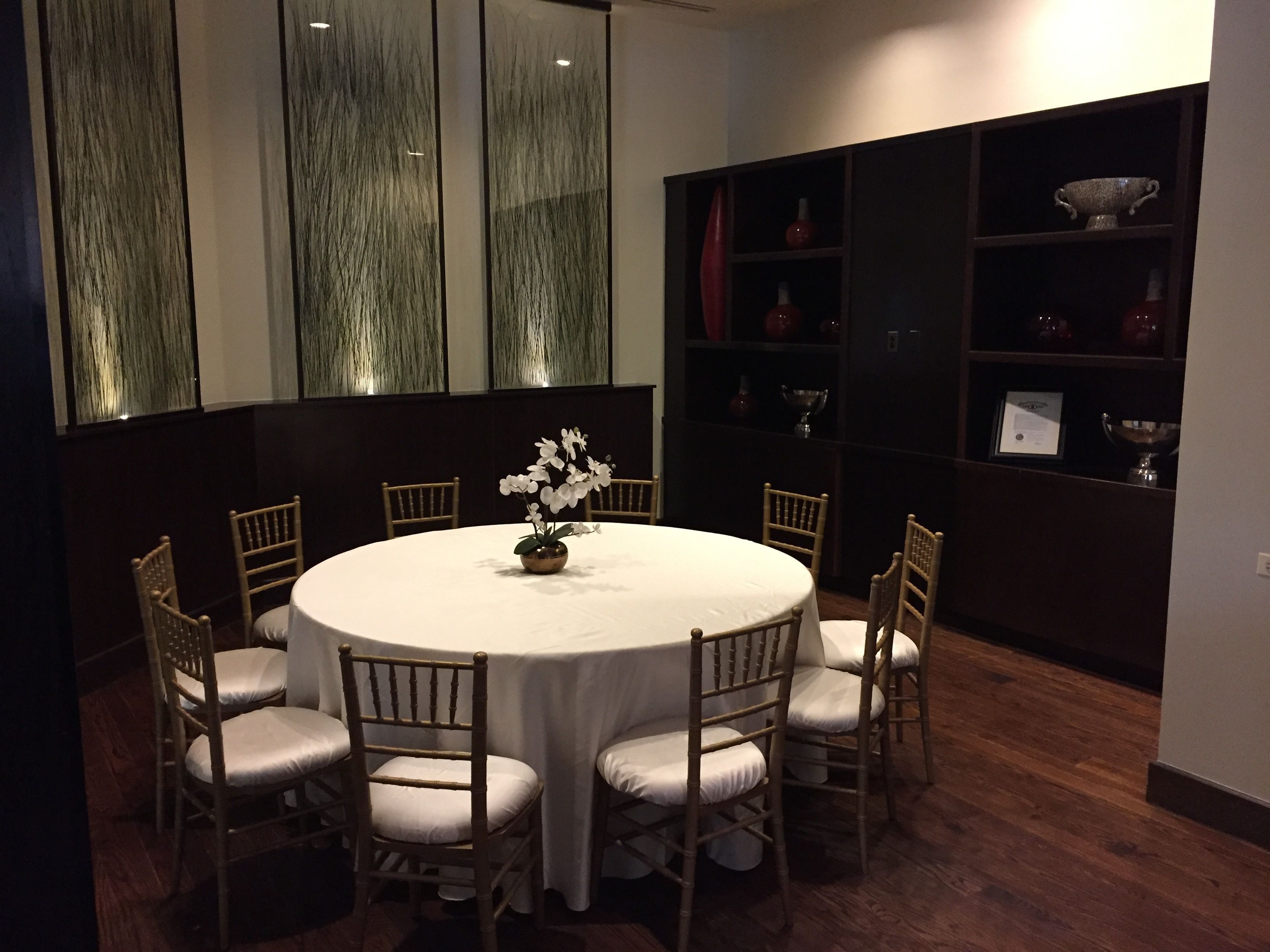 Una mesa redonda de comedor con mantel blanco, rodeada por seis sillas de madera con cojines blancos. Detrás de la mesa, hay una estantería de madera oscura con varios objetos decorativos.