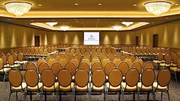 Un amplio salón de conferencias con filas de sillas, una pantalla de proyector mostrando el logotipo de Hilton y candelabros en el techo.