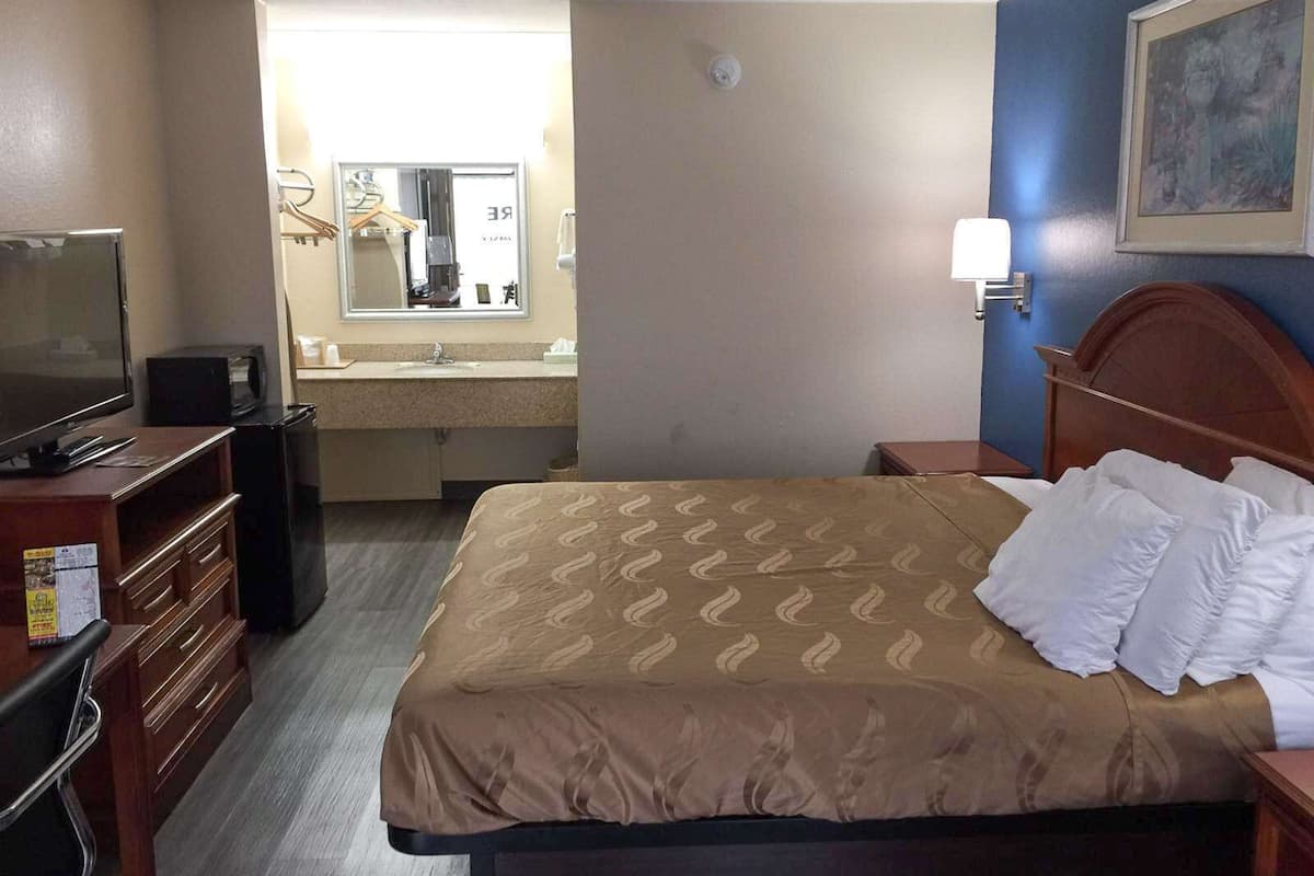 A hotel room with a bed, a television, a desk with drawers, a mirror, a sink, and a painting on the wall.