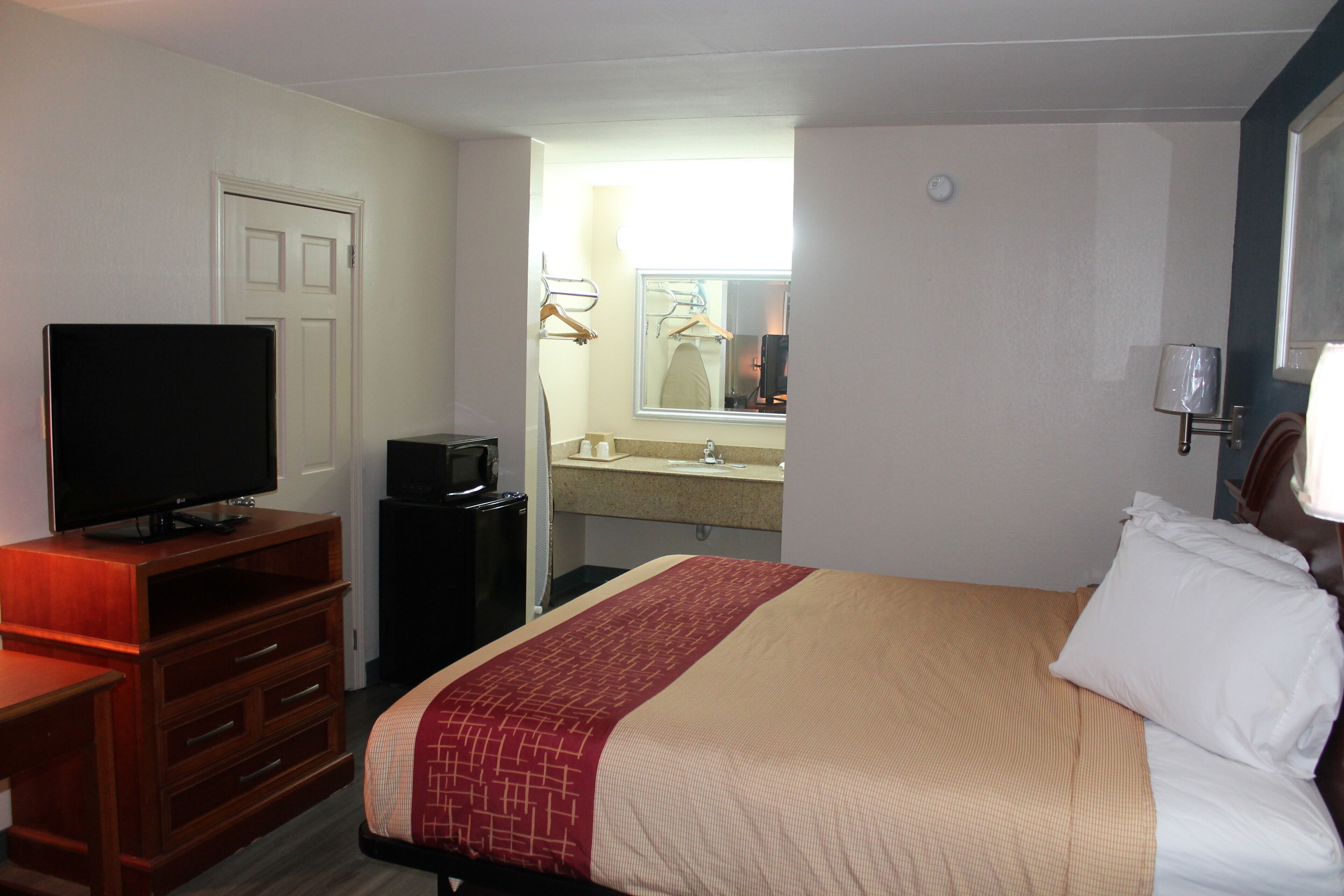 A hotel room with a bed, a TV on a wooden dresser, a bathroom with a sink and mirror, and a door.
