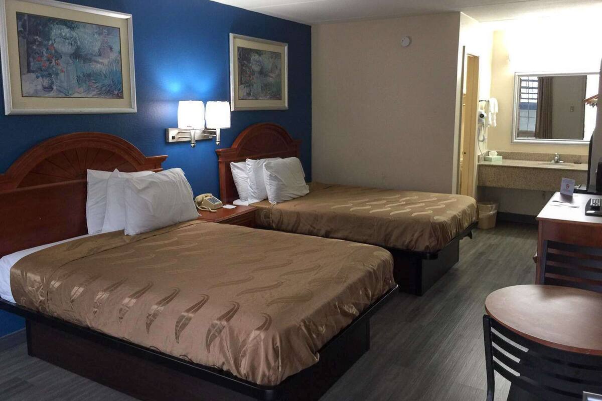 A hotel room with two beds, a desk, a chair, a mirror, and two wall-mounted lamps.