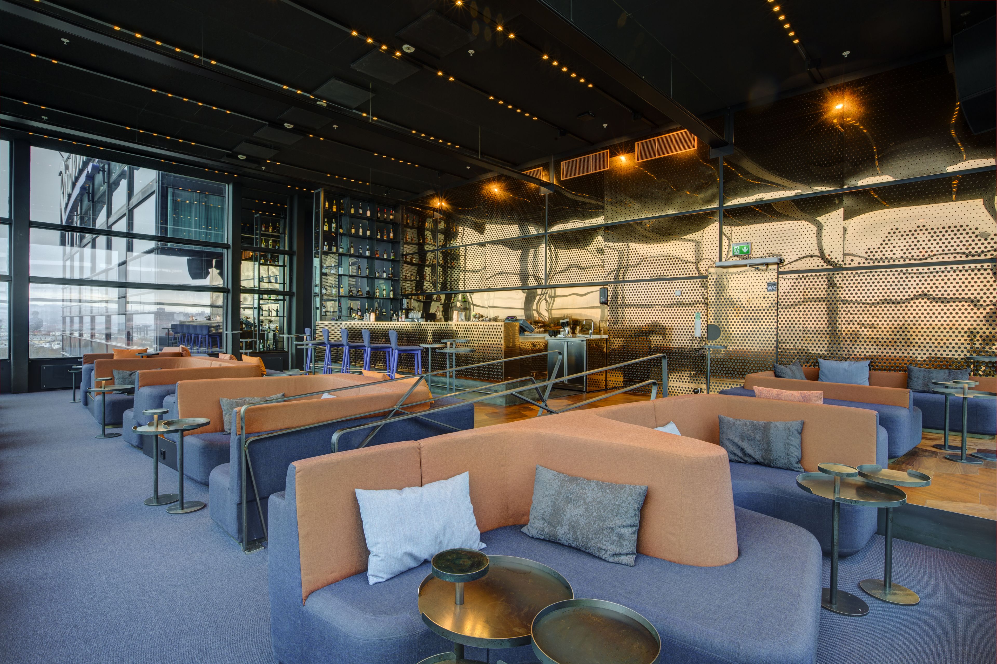 A modern lounge area with seating and tables, large windows, and a metal wall with a perforated design.