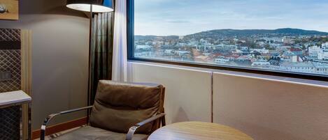 A hotel room with a leather chair, a small wooden table, a lamp, and a large window offering a city view.