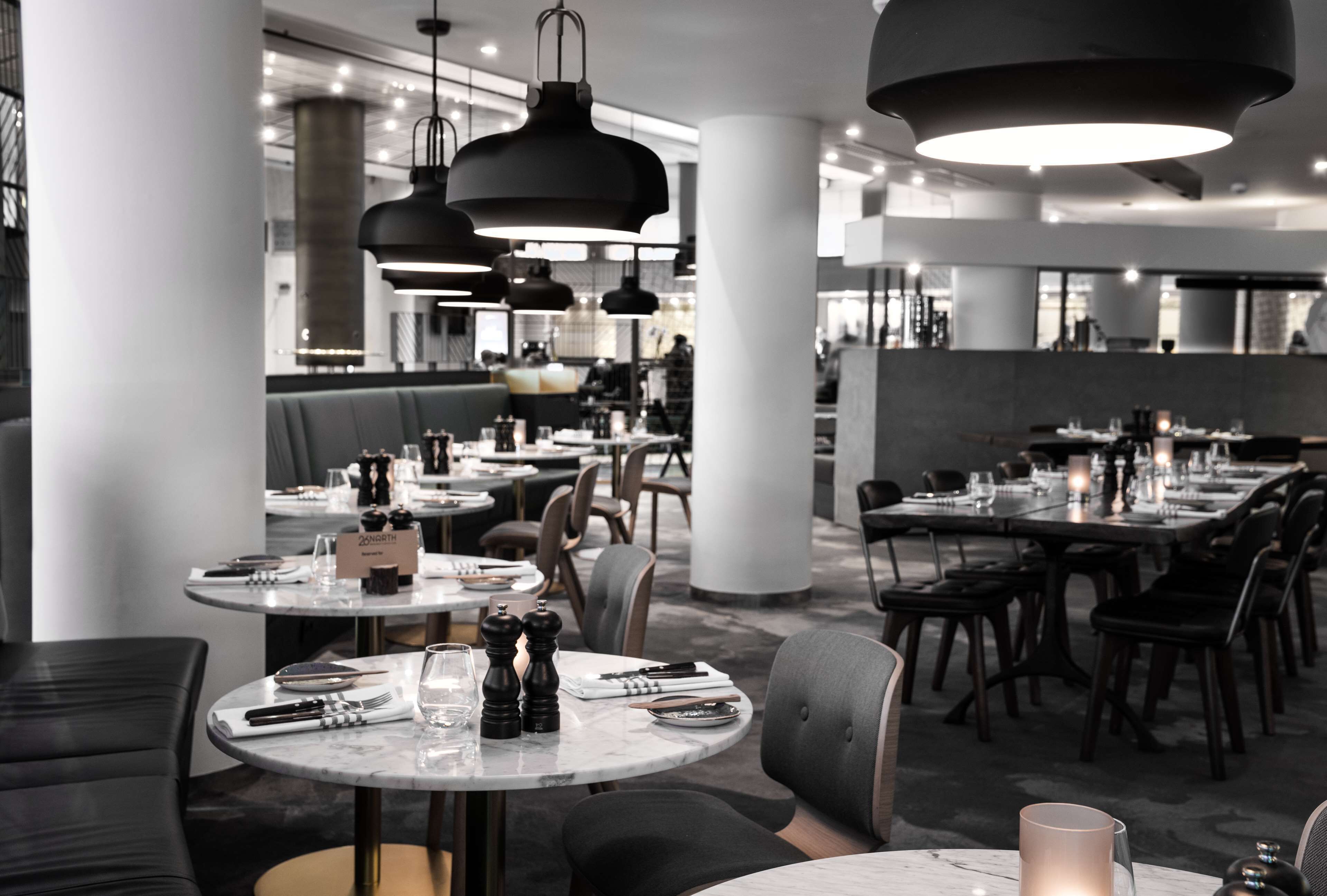 Un restaurant moderne avec des tables rondes, des luminaires suspendus noirs et une palette de couleurs neutres.