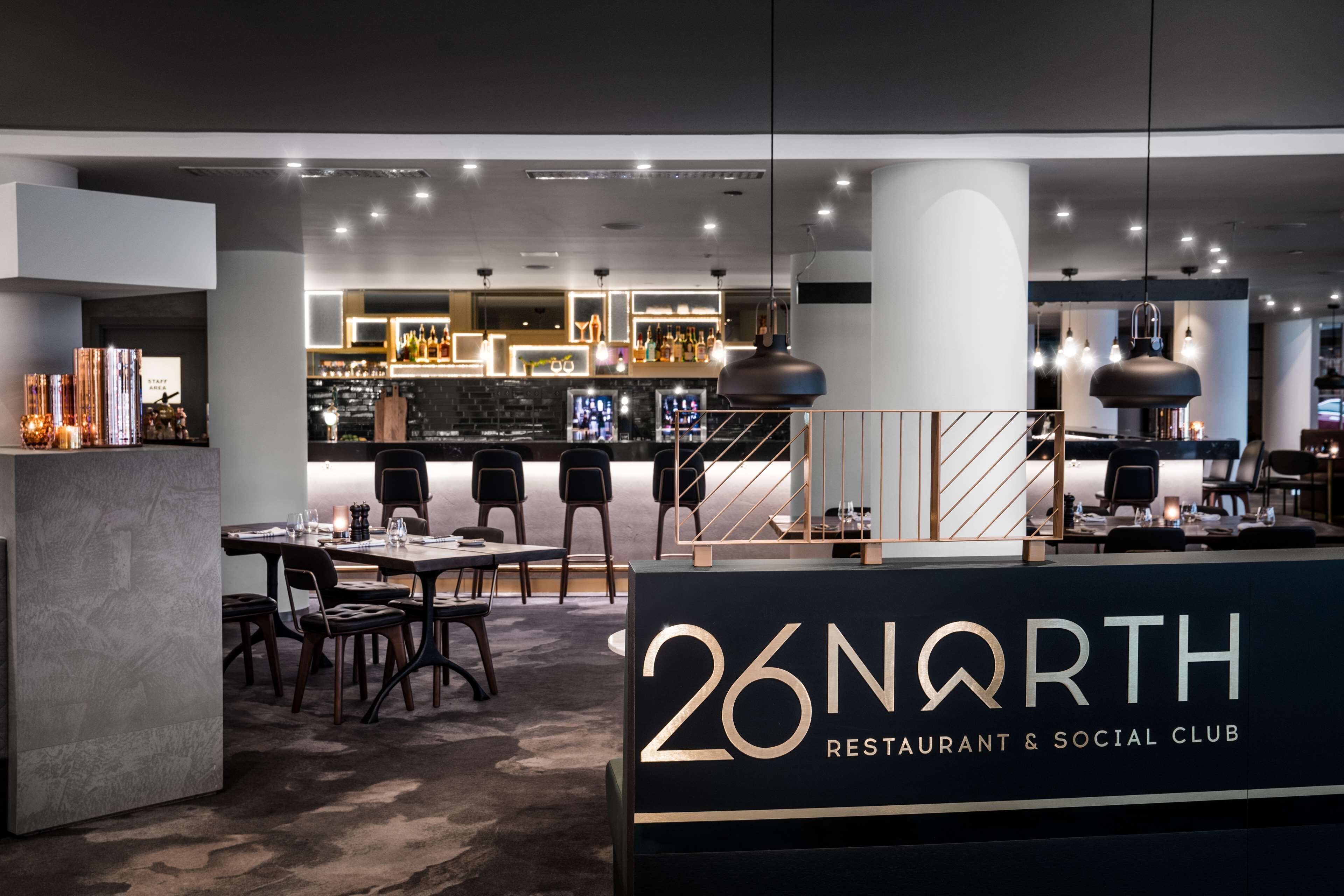 Ein modernes Restaurant-Interieur mit Barbereich, Sitzgelegenheiten und einem gut sichtbaren Schild mit der Aufschrift "26 NORTH RESTAURANT & SOCIAL CLUB".