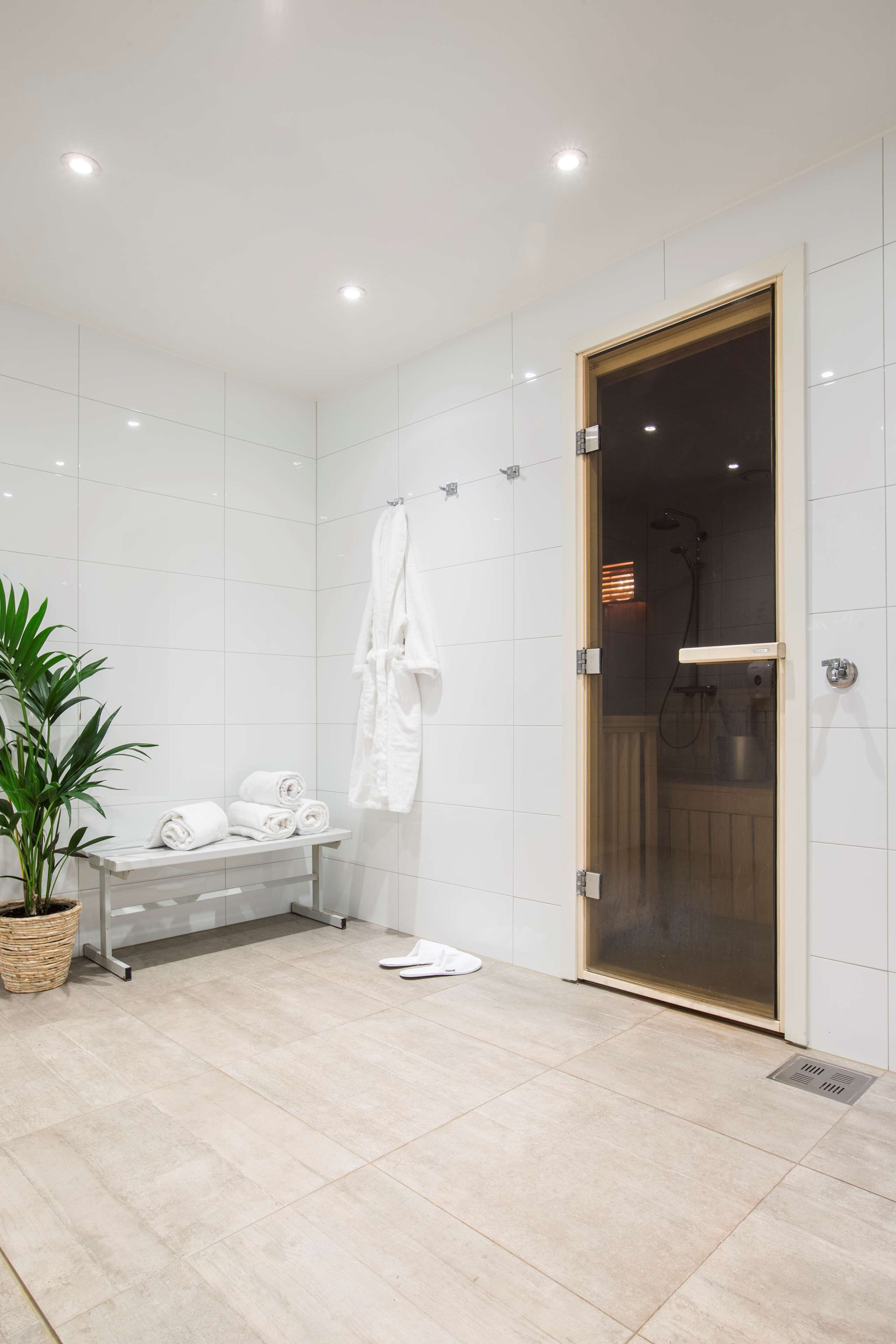 Un baño moderno con azulejos blancos, cabina de ducha con mampara de vidrio, toallero y planta en maceta.