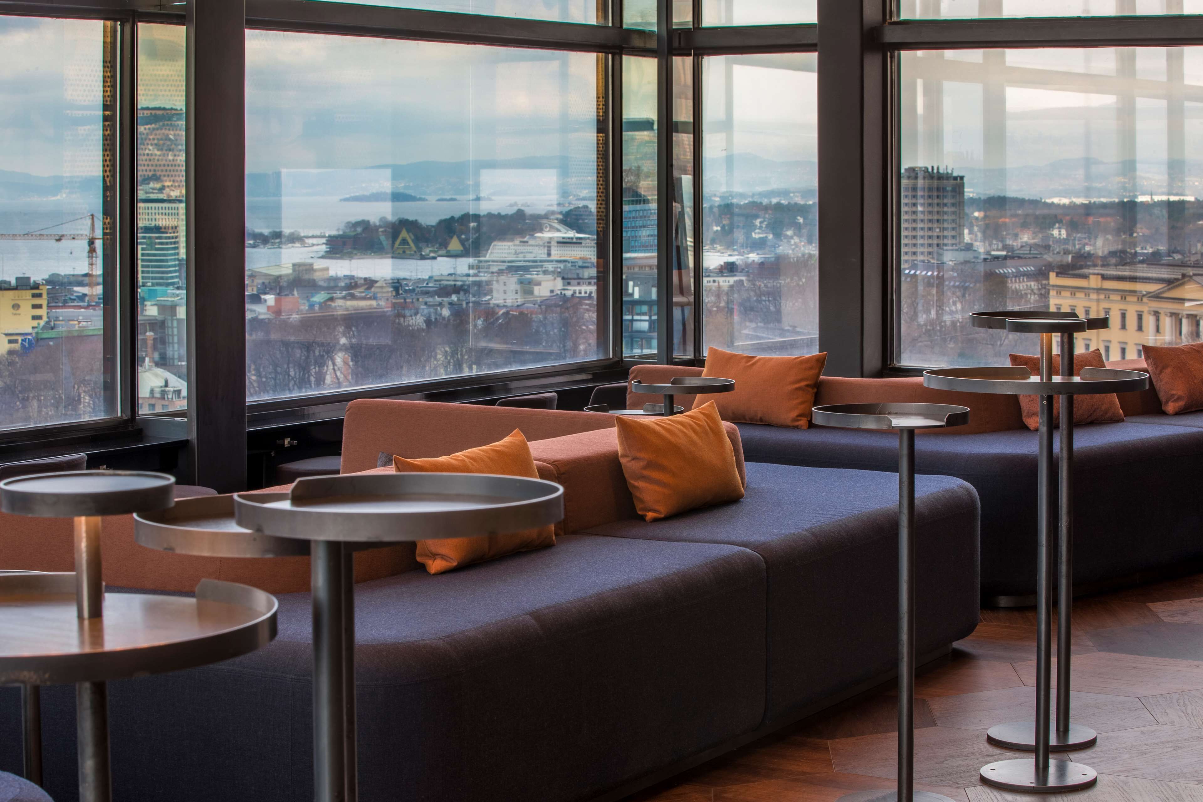 A modern lounge area with a large window offering a city view, cushioned seating, and round tables.