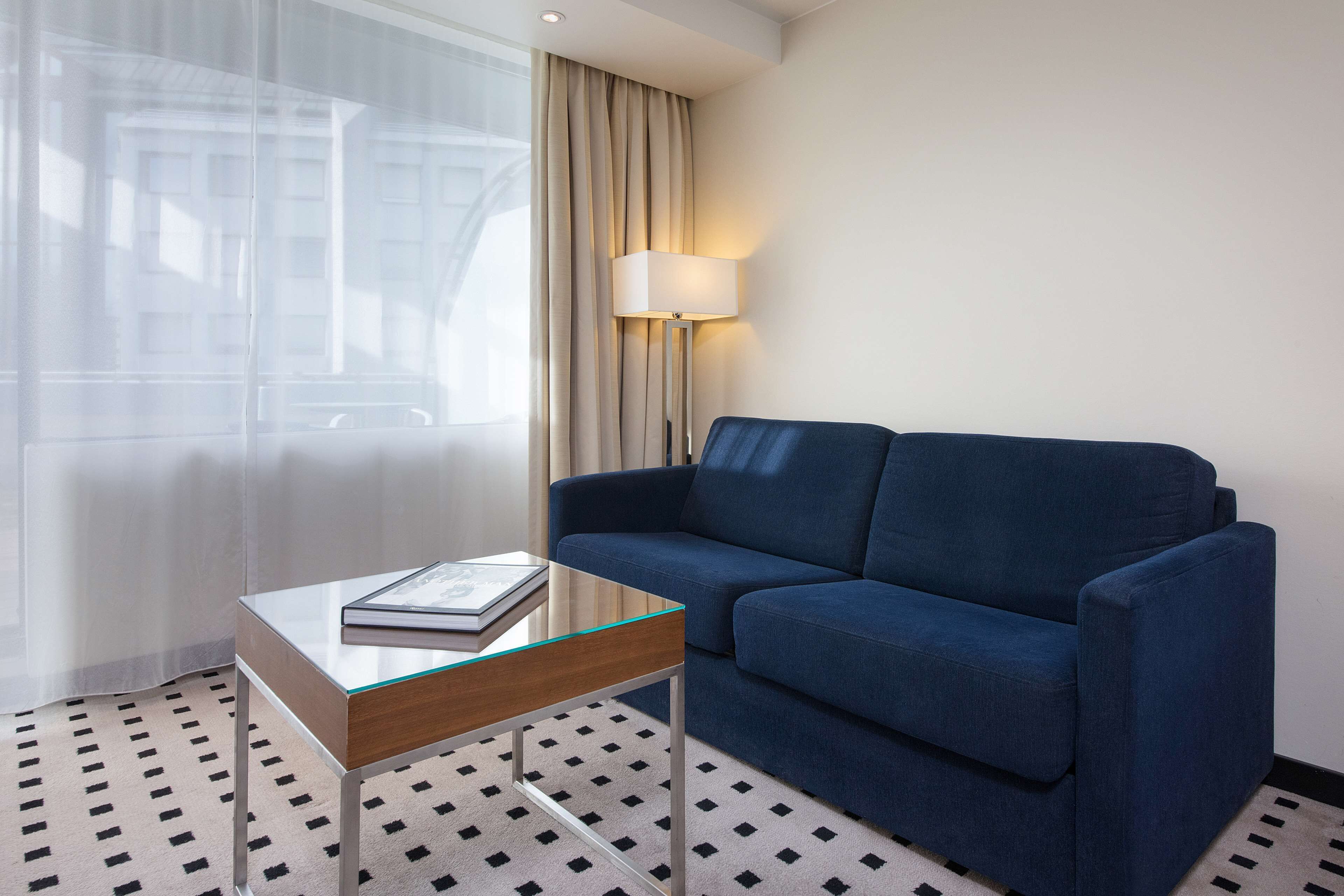 Una habitación de hotel moderna con un sofá azul, una mesa de centro de vidrio, una lámpara y una alfombra estampada.