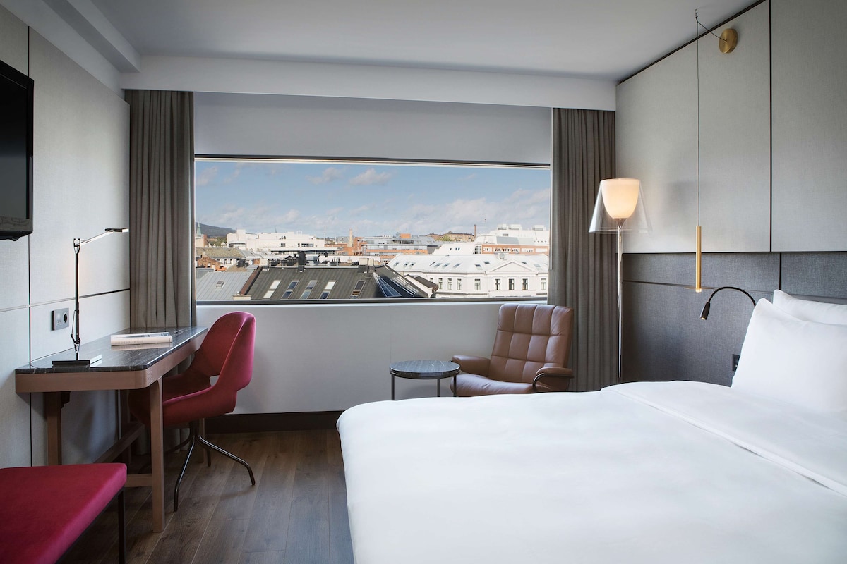 A hotel room with a large bed, a desk with a chair, a small table, and a view of the cityscape through a large window.
