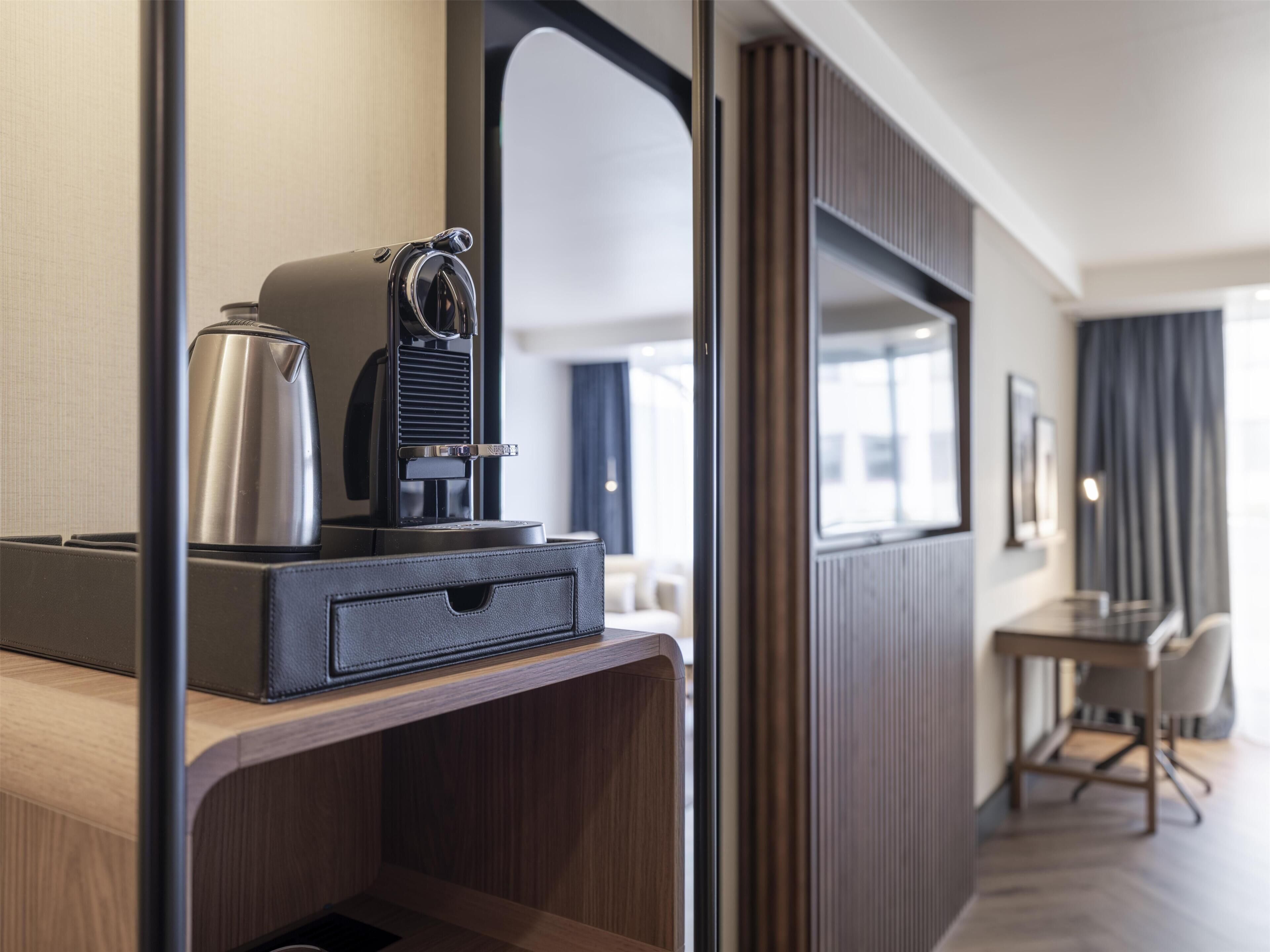 A hotel room with a wooden desk, a coffee maker, a mirror, and a view of the hallway.