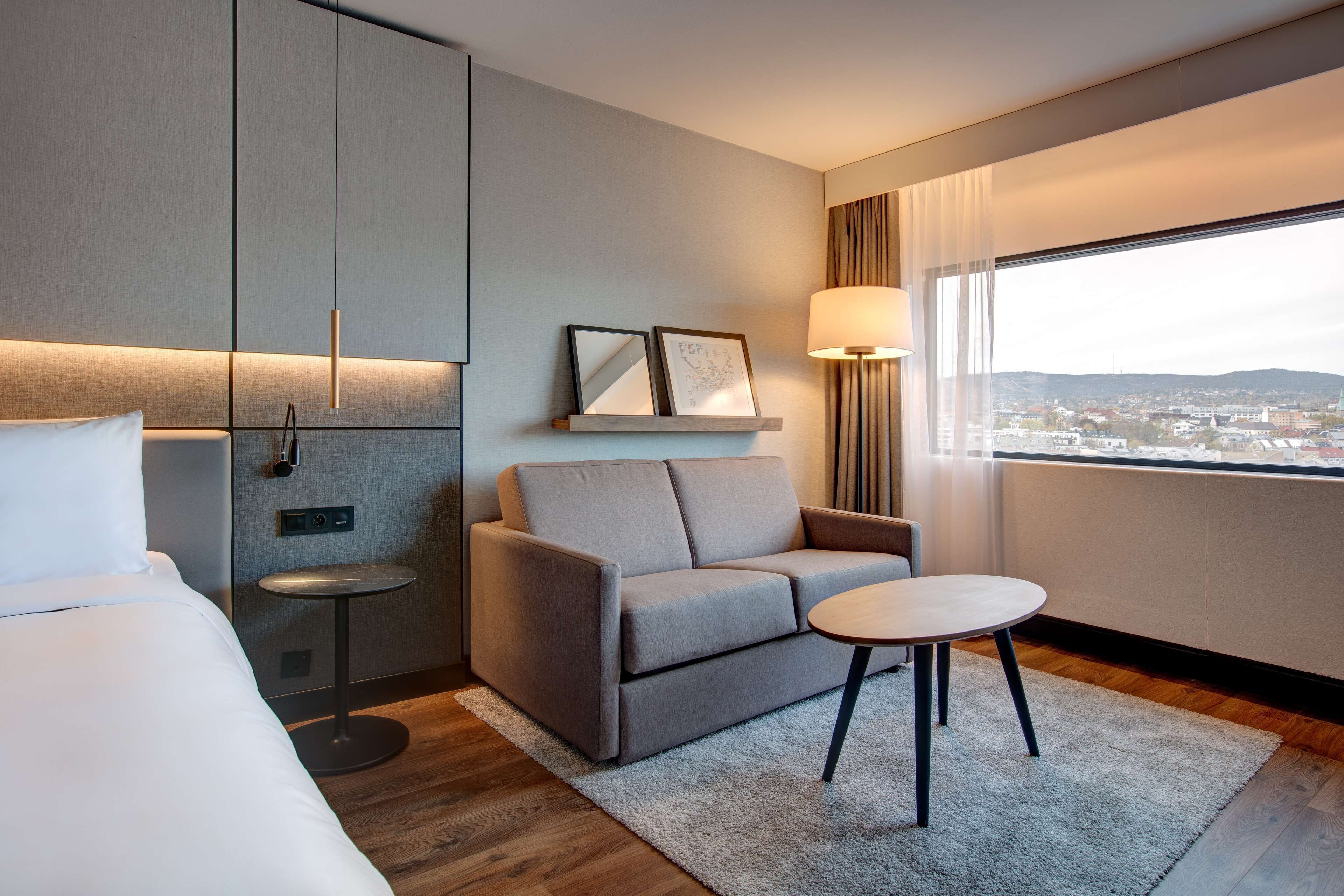 Habitación de hotel con cama, sofá, mesita, lámpara y ventana con vista a la ciudad.