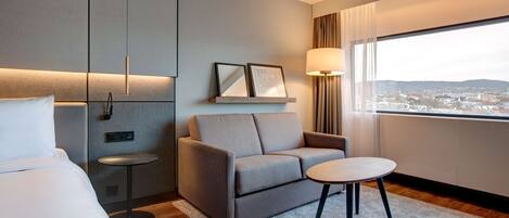 Habitación de hotel con cama, sofá, mesita, lámpara y ventana con vista a la ciudad.