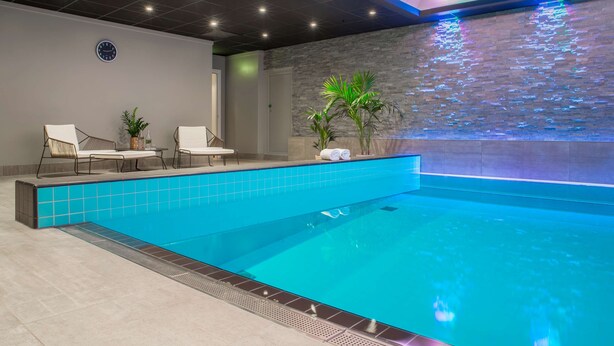 Un área de piscina interior moderna con una piscina azul cristalina, dos sillones de descanso, una mesita y un reloj fijado a la pared.