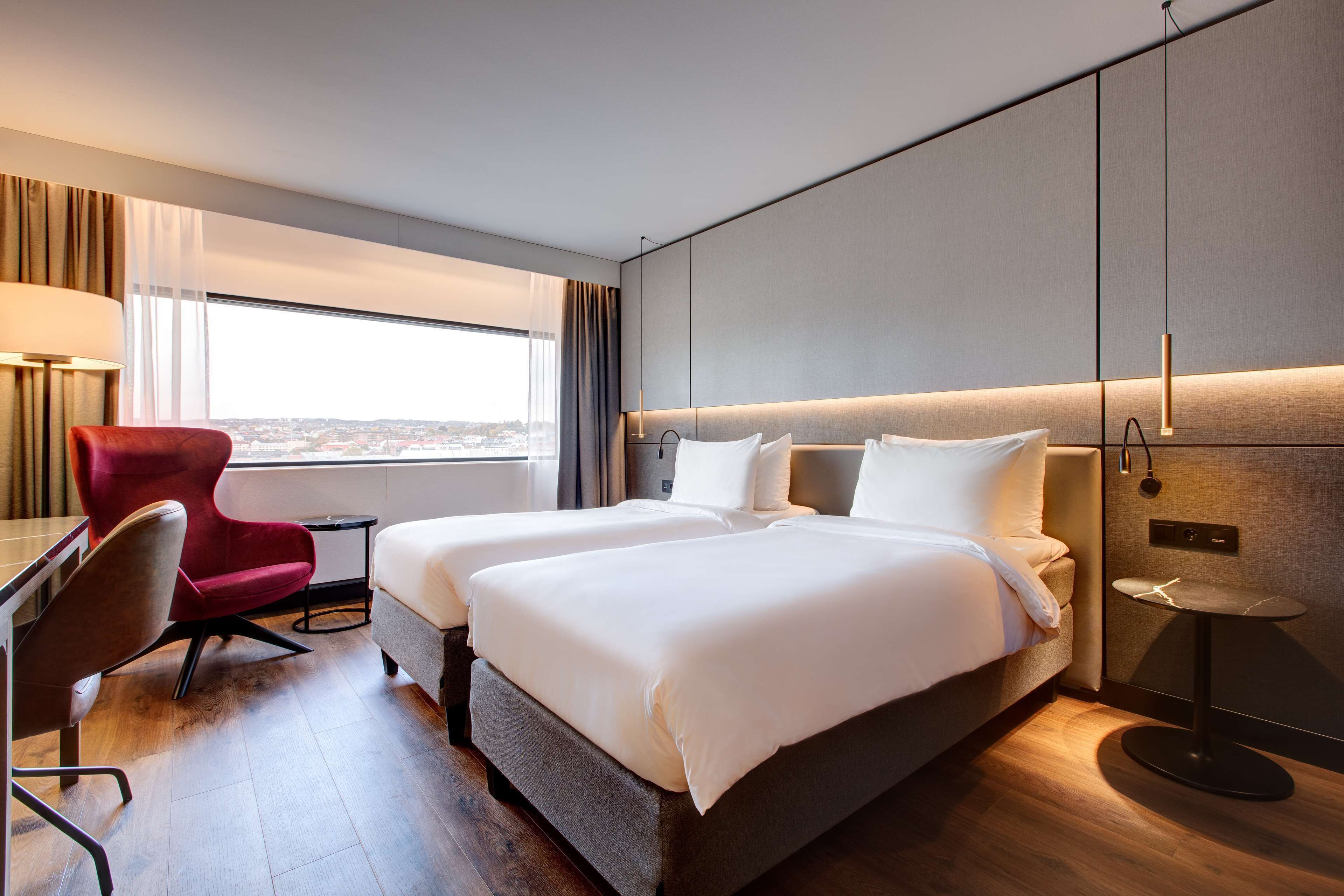 Habitación de hotel con dos camas, una silla roja, un escritorio y vistas a la ciudad.