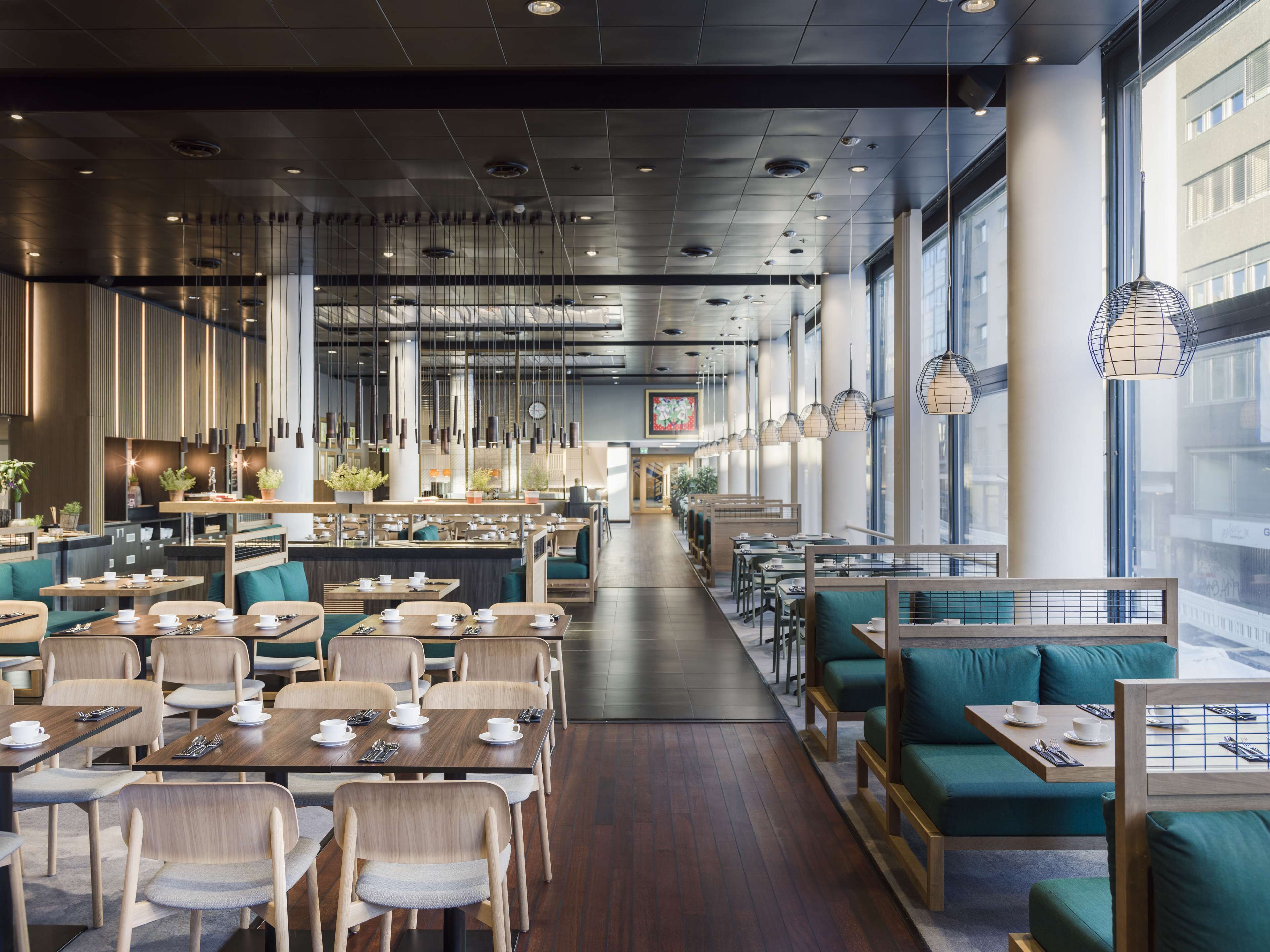 A modern restaurant with wooden tables and green cushioned seating, large windows, and hanging pendant lights.