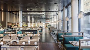 A modern restaurant with wooden tables and green cushioned seating, large windows, and hanging pendant lights.