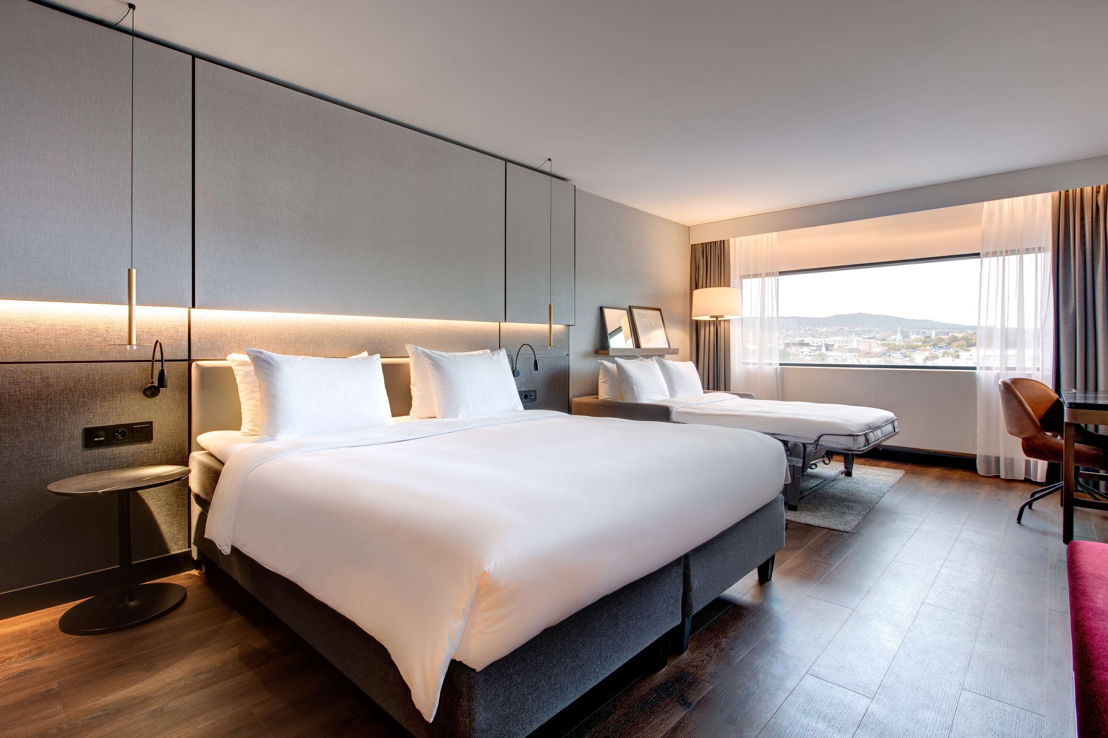 Een moderne hotelkamer met een groot bed, een kleiner bed, een bureau en een stoel. Er is een raam met uitzicht op de stad.