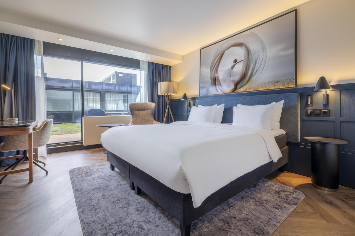 A modern hotel room with a large bed, a desk, a chair, and a view of the outside.
