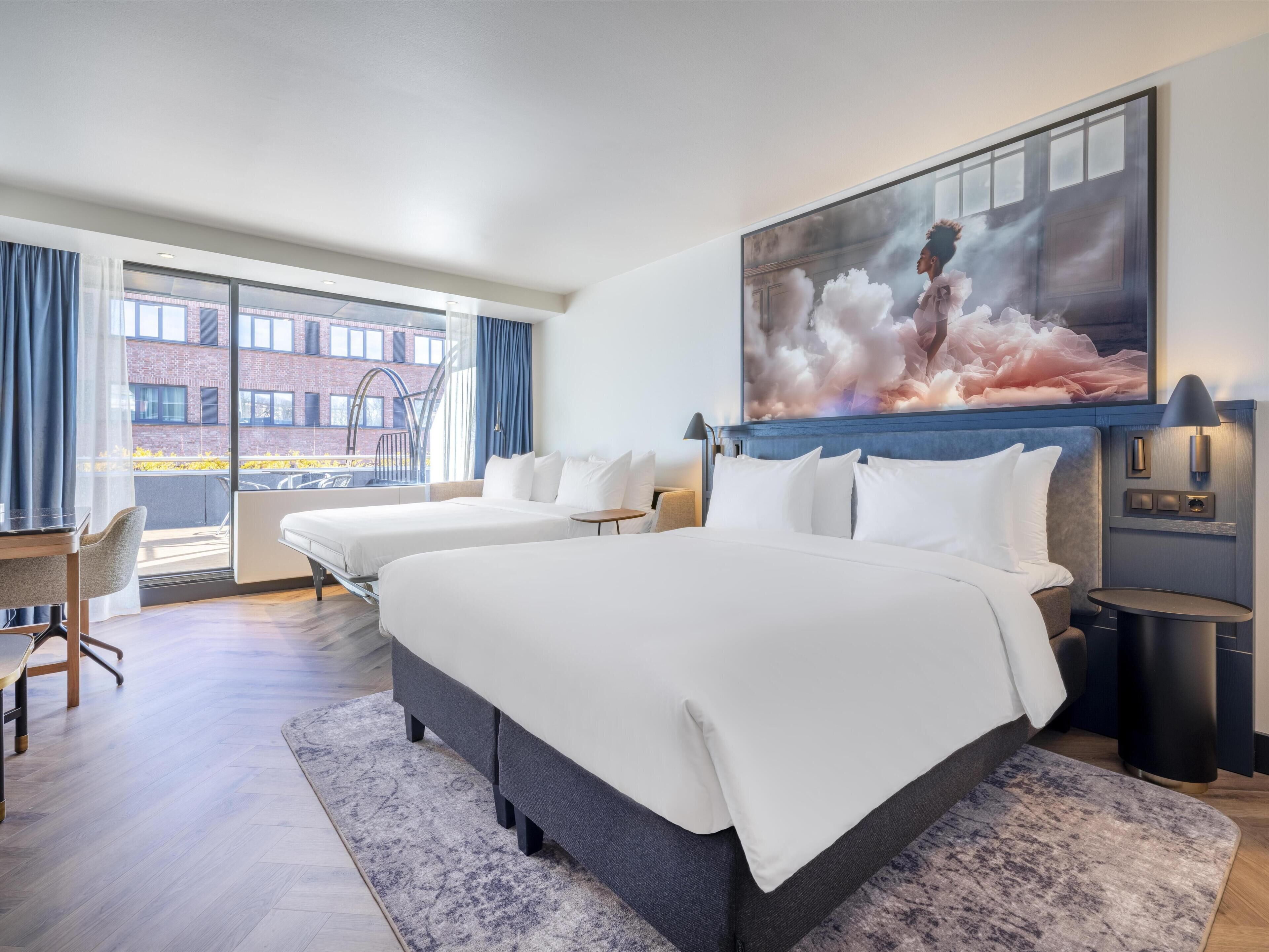 Habitación de hotel con una cama grande, un escritorio y una silla. Se ve una ventana con vistas a un edificio y un balcón.