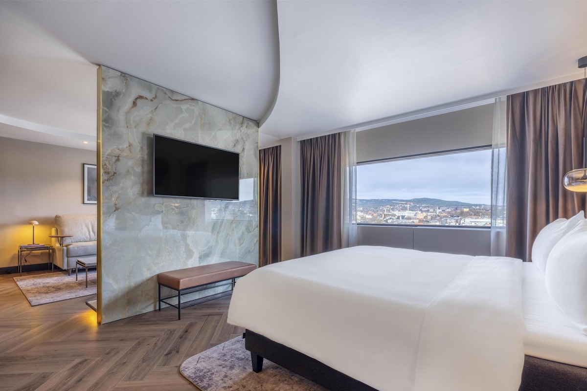 A modern hotel room with a large bed, a flat-screen TV mounted on a marble wall, a bench, a small table with a lamp, and a city view through large windows.