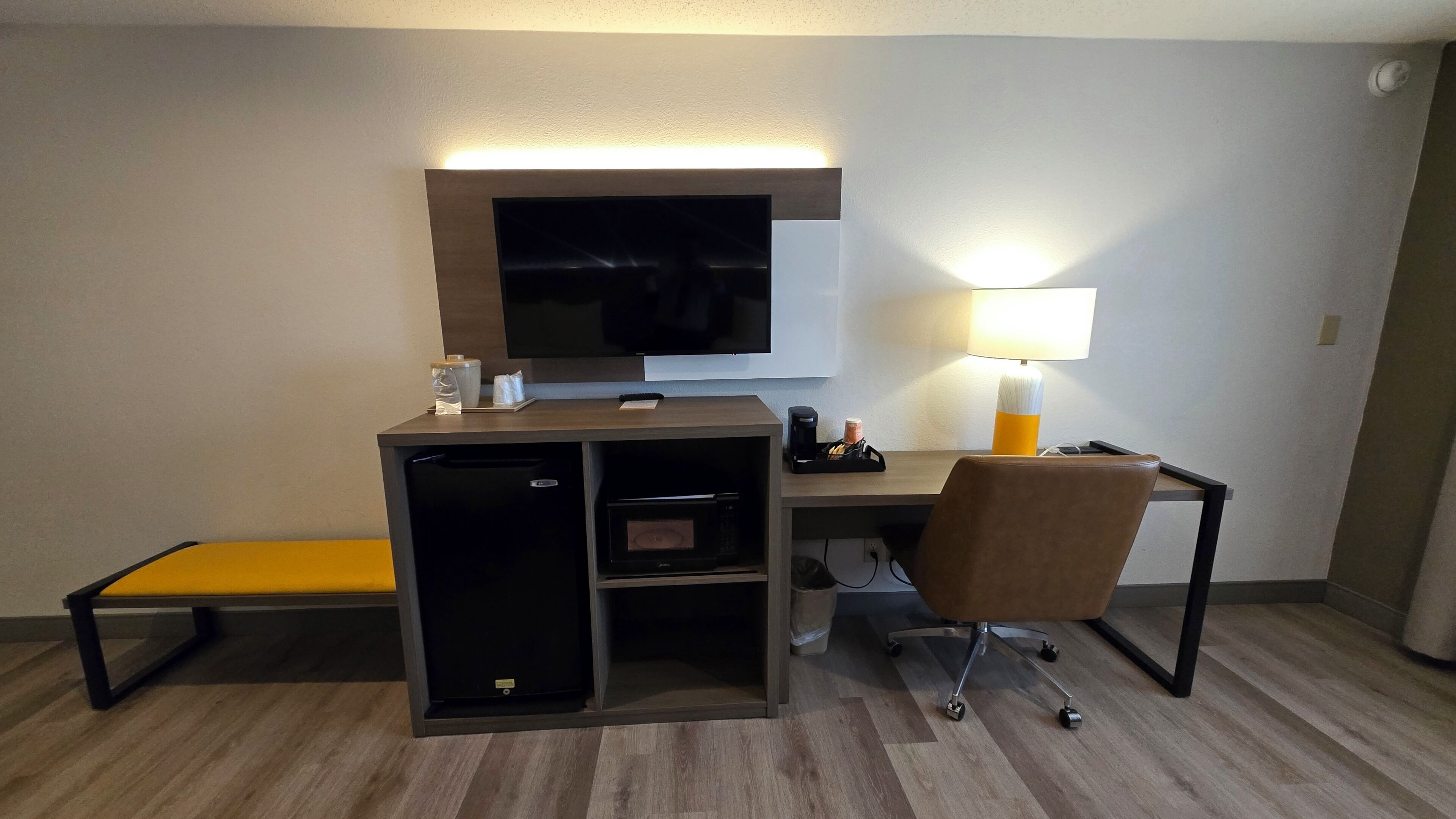 A hotel room with a desk, a television, a lamp, a microwave, a refrigerator, and a bench.
