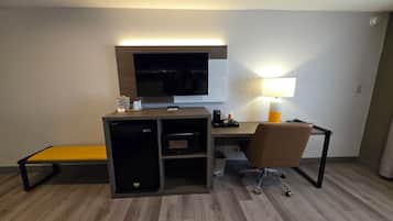 Habitación de hotel con escritorio, televisor, lámpara, microondas, refrigerador y una banca.