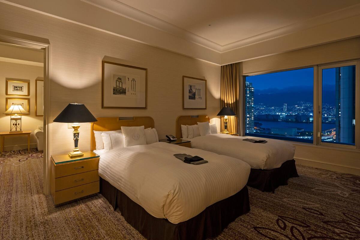 Una habitación de hotel con dos camas, una mesita de noche con lámpara, un escritorio con lámpara y un ventanal con vista a la ciudad.