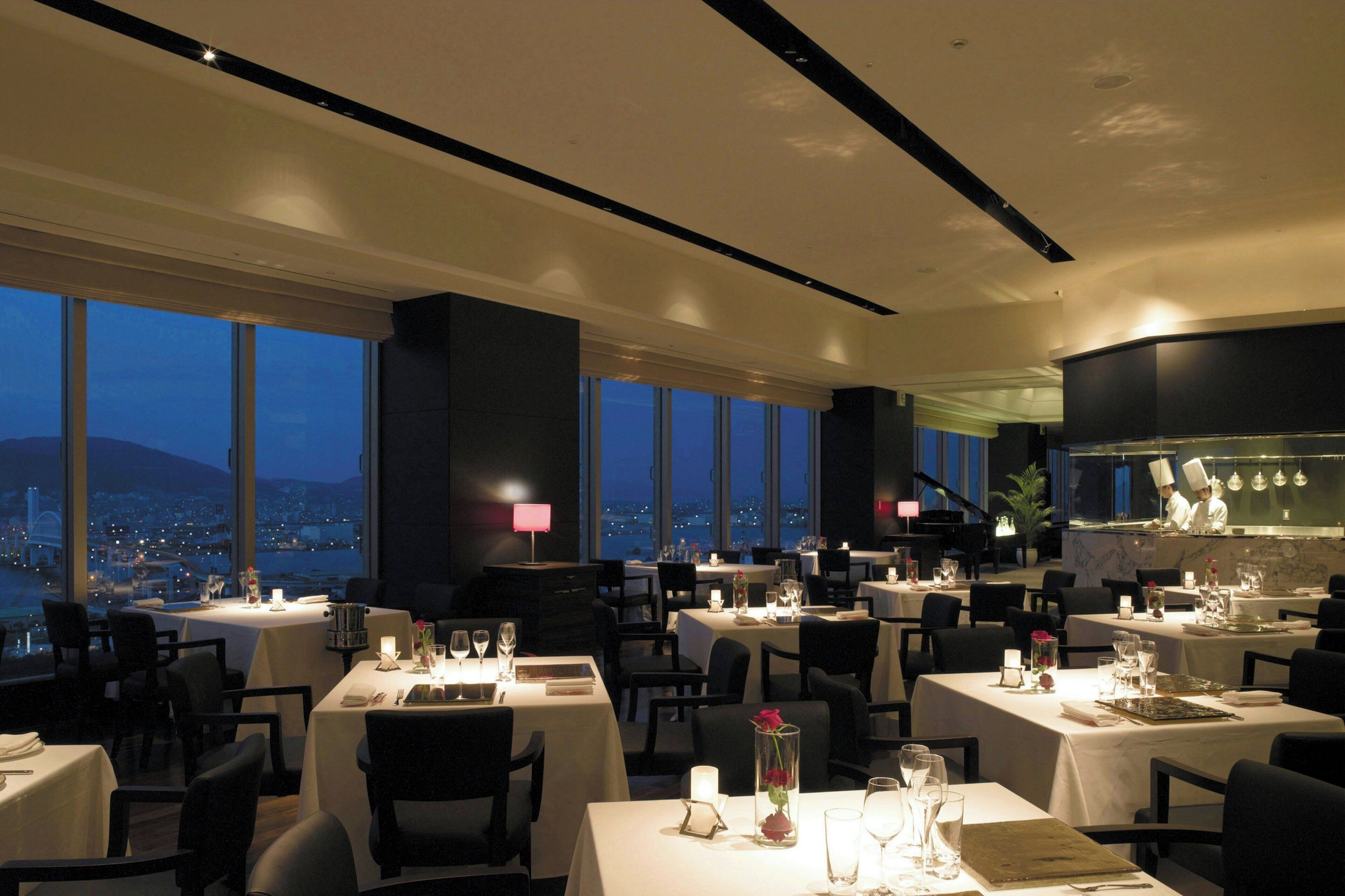 A restaurant with tables set for dining, a chef in the background, and a cityscape view through large windows.