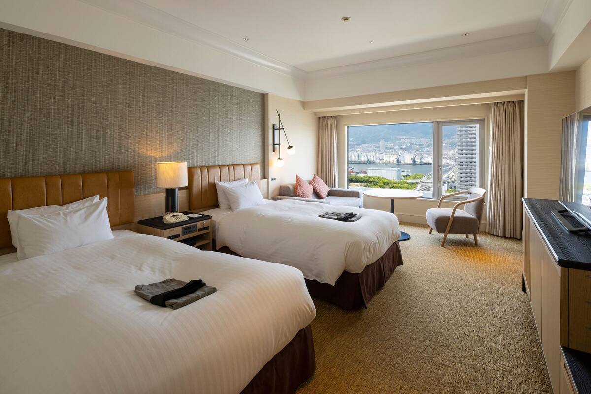 Habitación de hotel con dos camas, un escritorio, una silla y un ventanal con vistas a la ciudad.