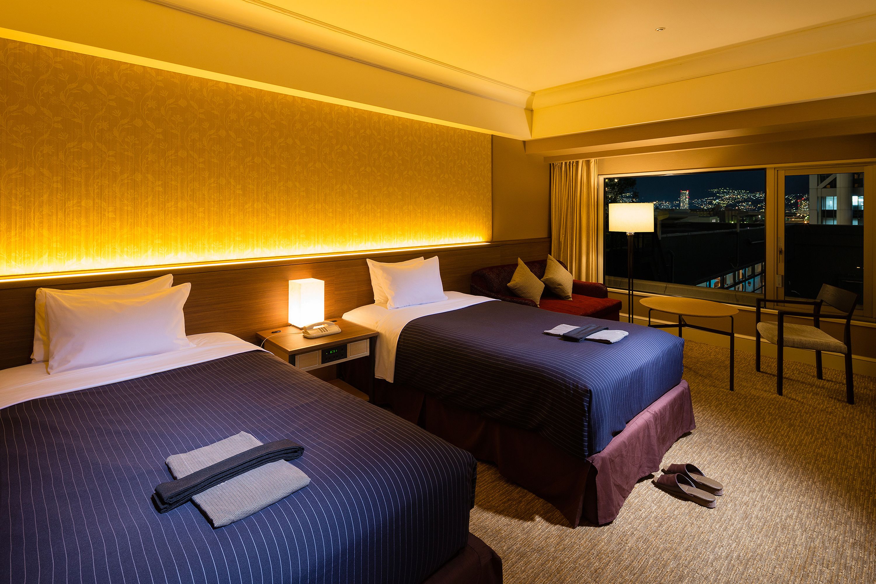 A hotel room with two beds, a nightstand with a lamp, a chair, and a view of the city at night.