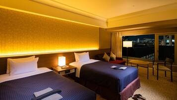 A hotel room with two beds, a nightstand with a lamp, a chair, and a view of the city at night.