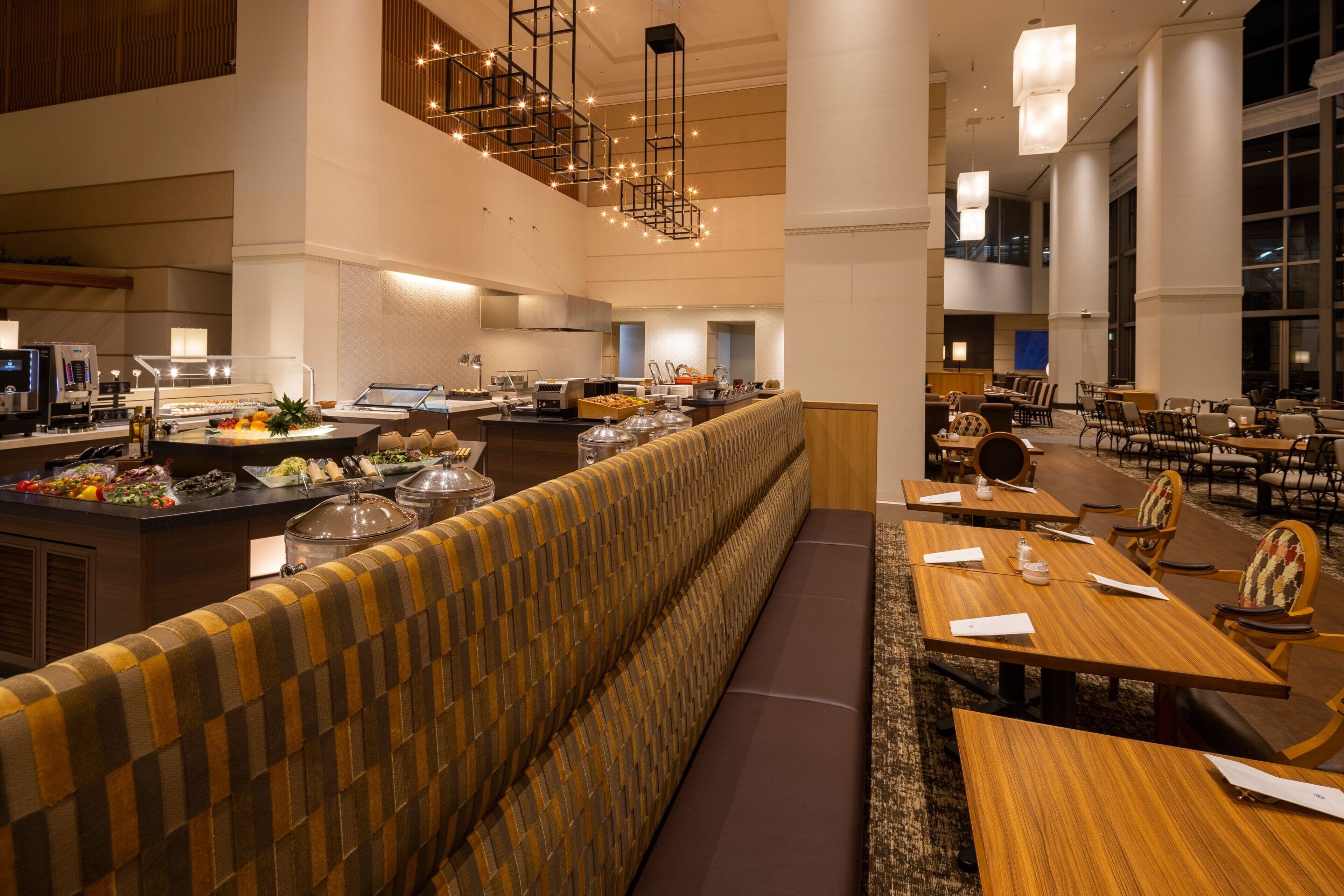 A spacious hotel dining area with a long counter, seating arrangements, and a high ceiling with hanging lights.