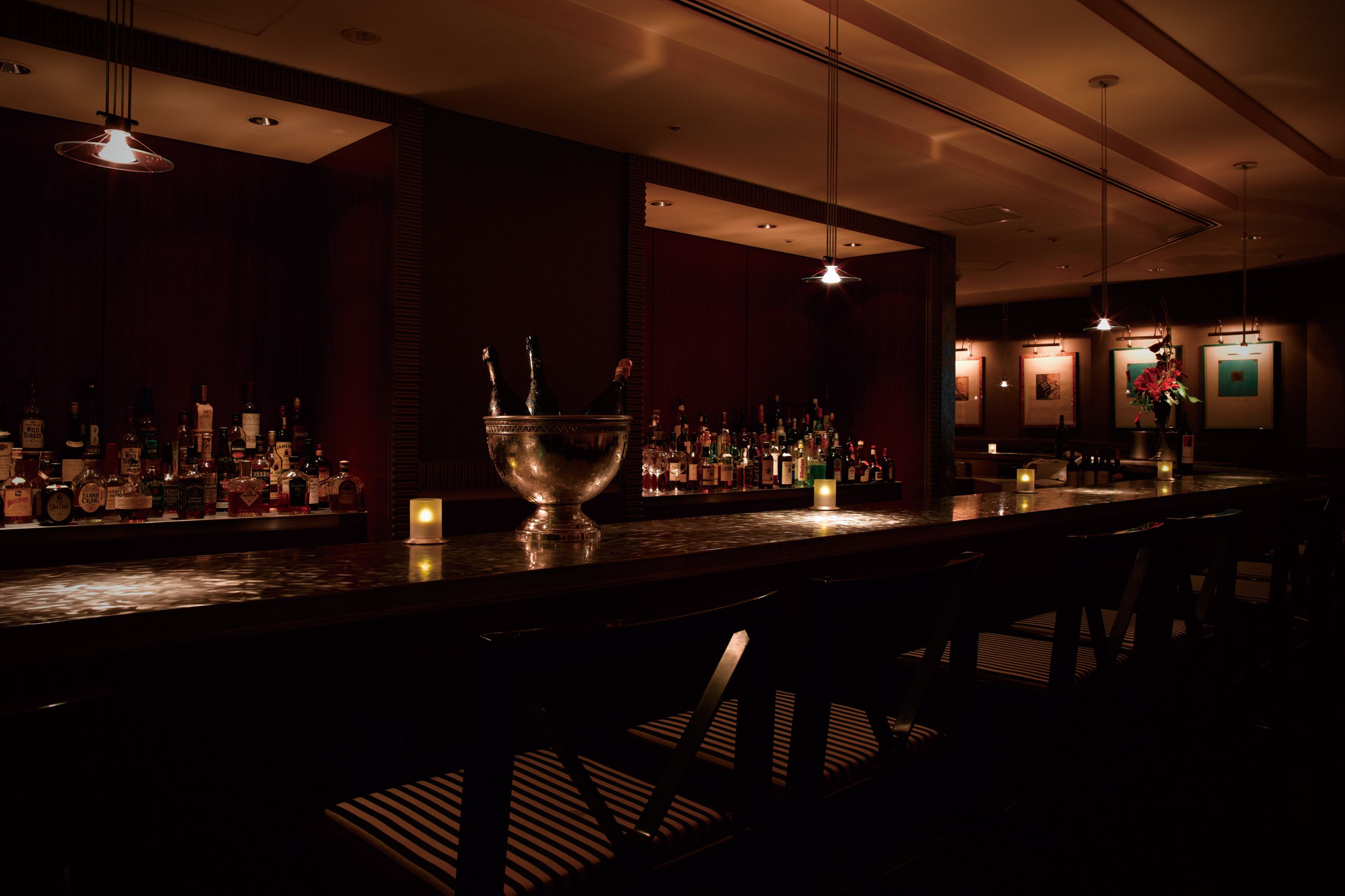 Zona de bar tenuemente iluminada con una barra pulida, varias botellas de licores y un recipiente decorativo grande.