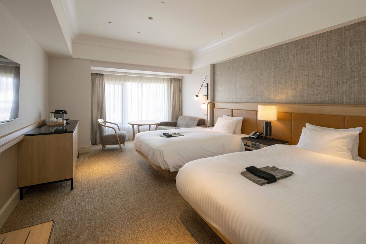 Habitación de hotel con dos camas, un escritorio, una silla, una lámpara y un ventanal con cortinas.