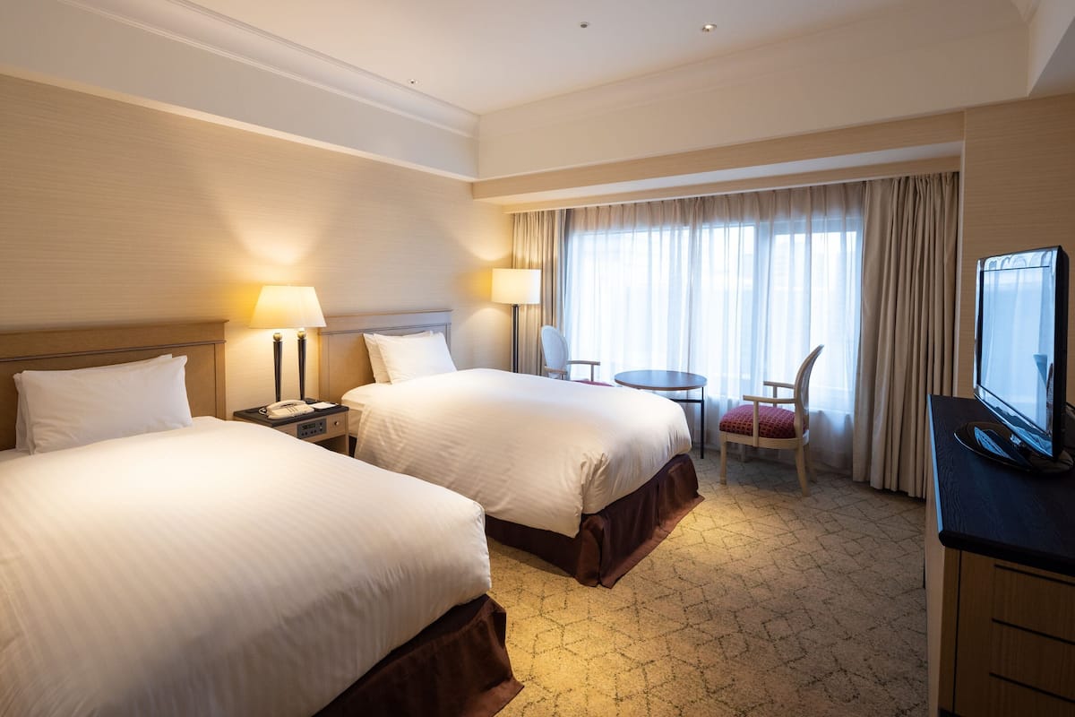 Habitación de hotel con dos camas, un escritorio con silla, televisión y ventana con cortinas.