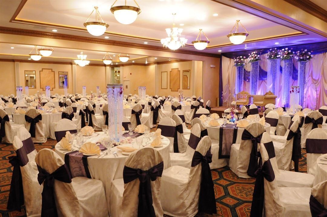 Une salle de banquet avec des tables rondes, des chaises ornées de rubans, des lustres et une scène décorée de draperies.